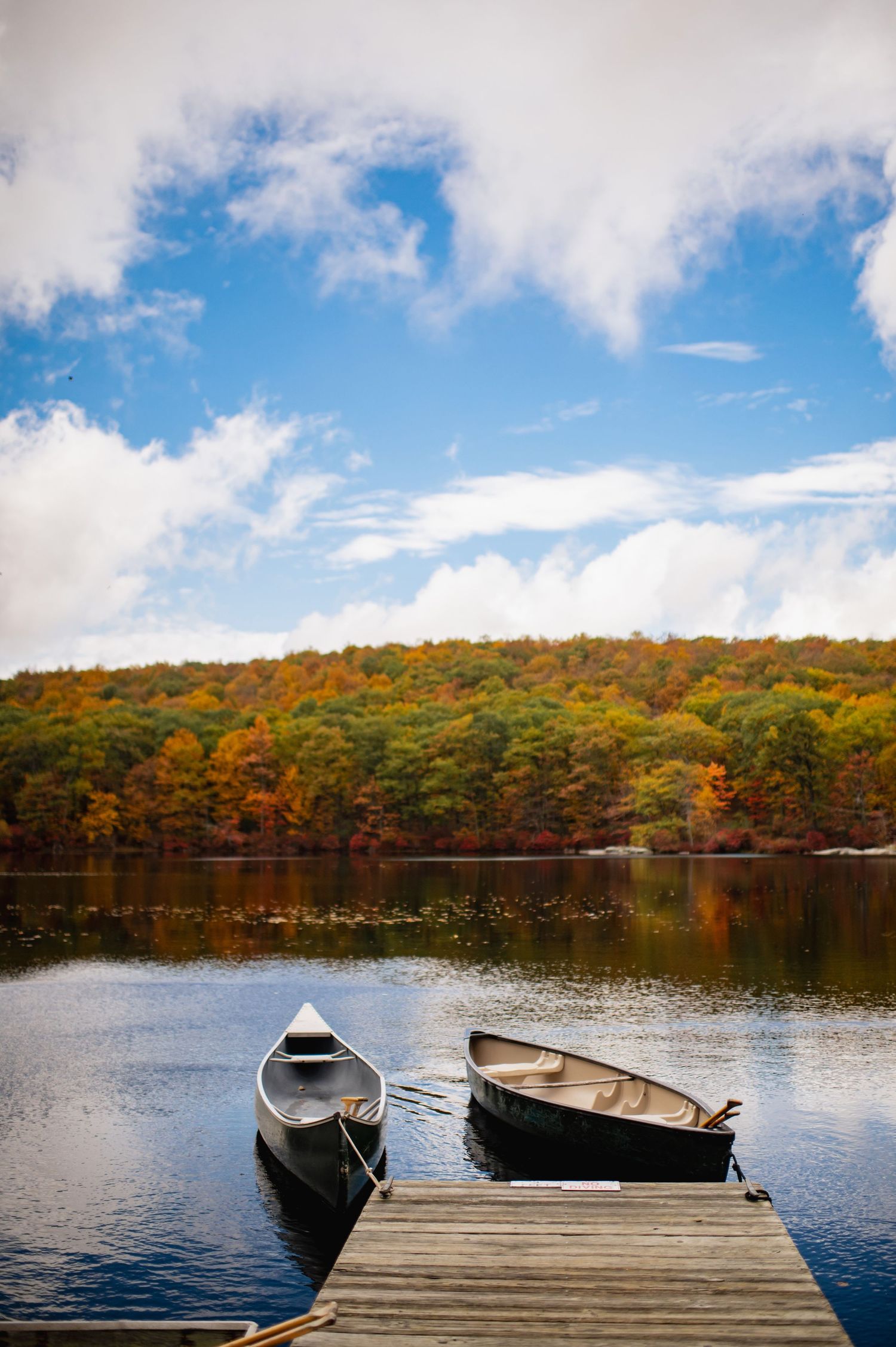 Cedar Lakes venue in upstate NY; Two wooden boats tied to a dock on a calm lake surrounded by autumn foliage and white puffy clouds.