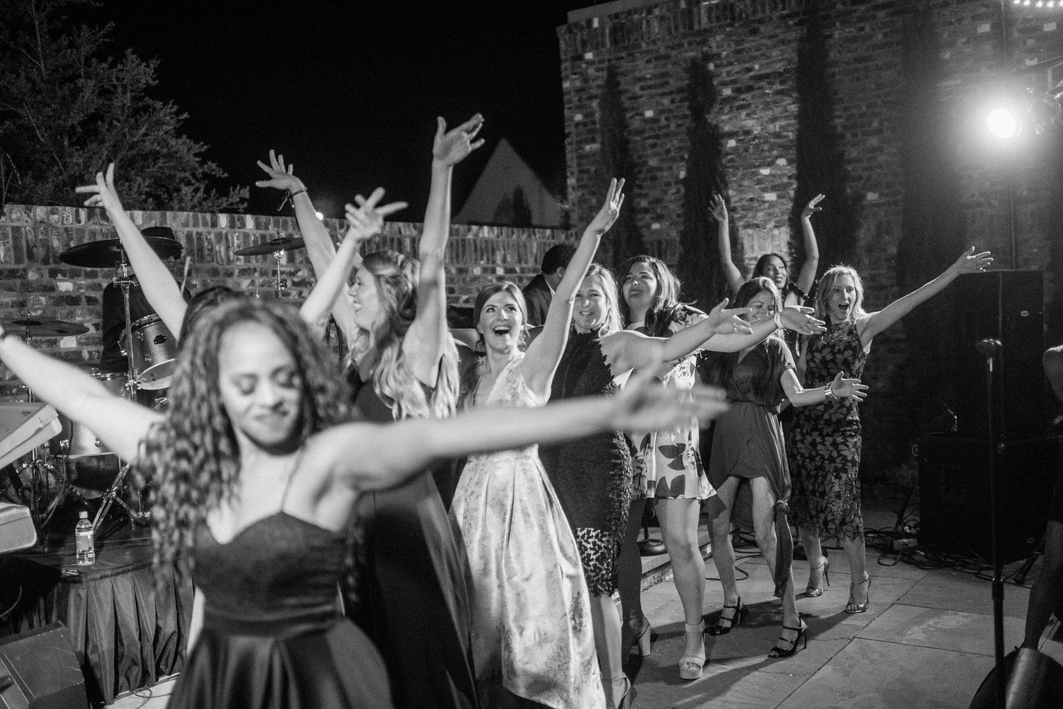 Wedding reception guests dance enthusiastically with raised arms on a crowded dance floor at night.
