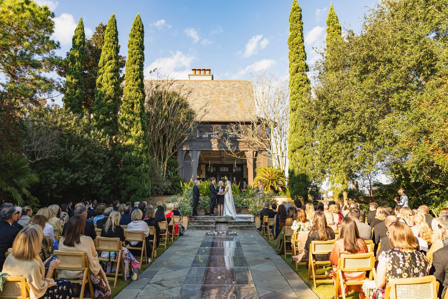 Outdoor wedding ceremony setup with wooden chairs and tall cypress trees lining a stone pathway in a garden setting.