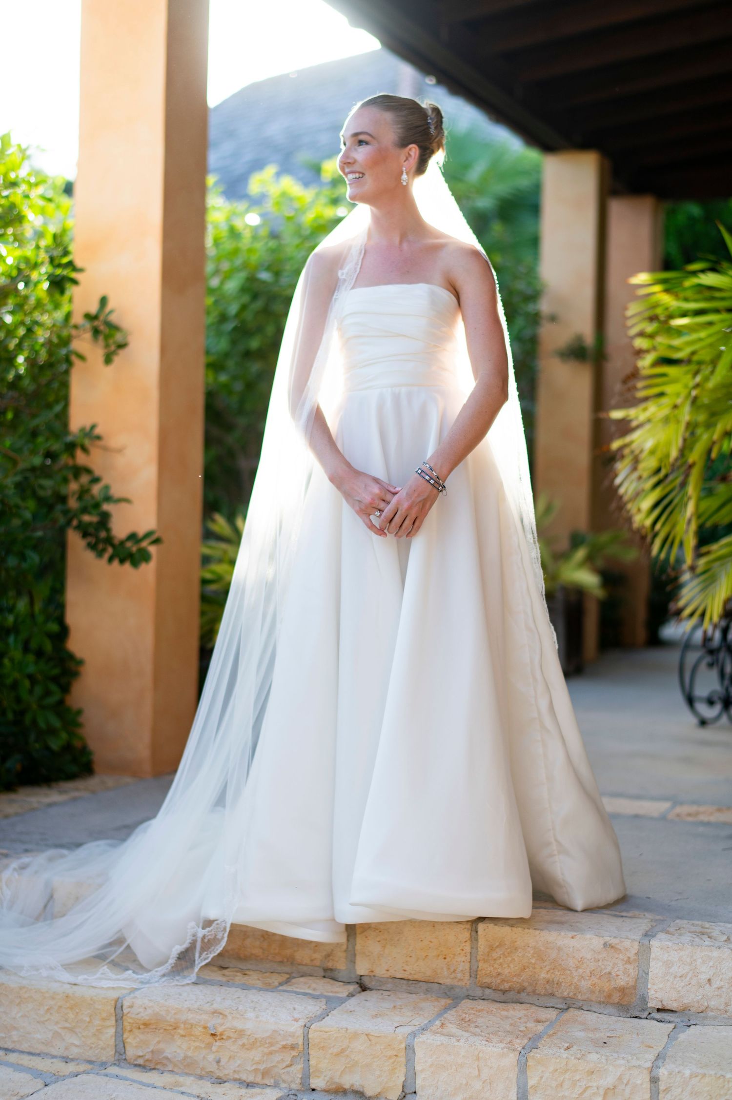 Elegant white wedding dress with flowing veil photographed against stone columns and tropical foliage.