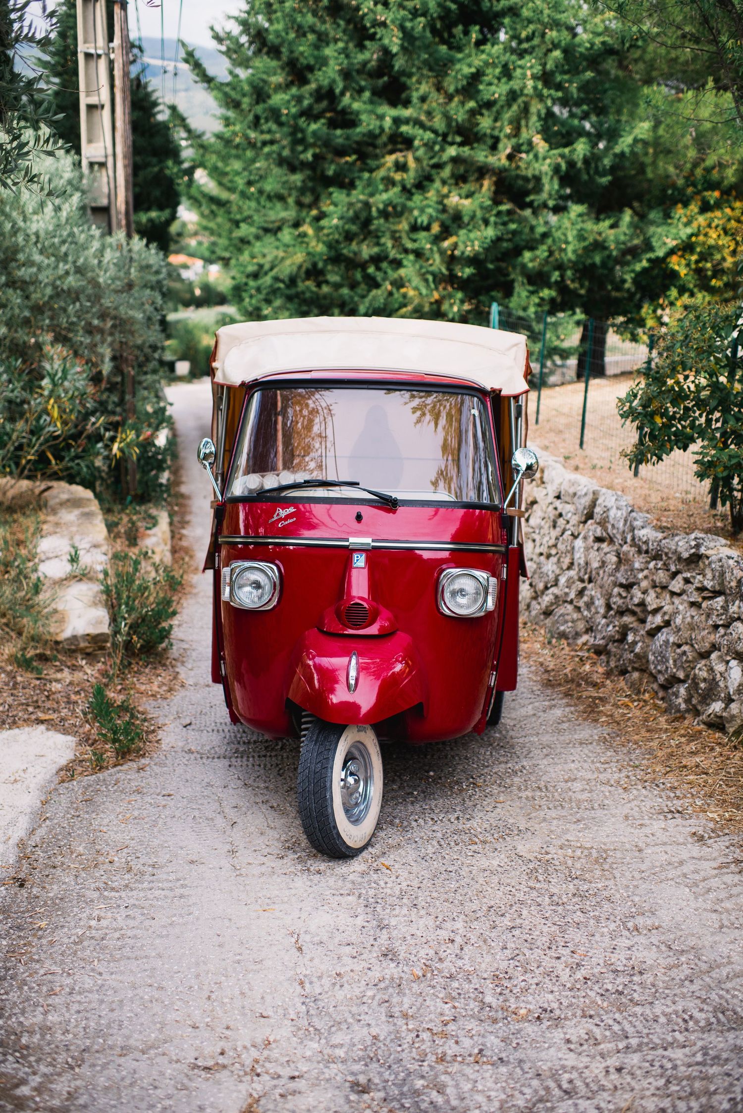 Vintage red three-wheeled vehicle parked on a rustic gravel path lined with stone walls.