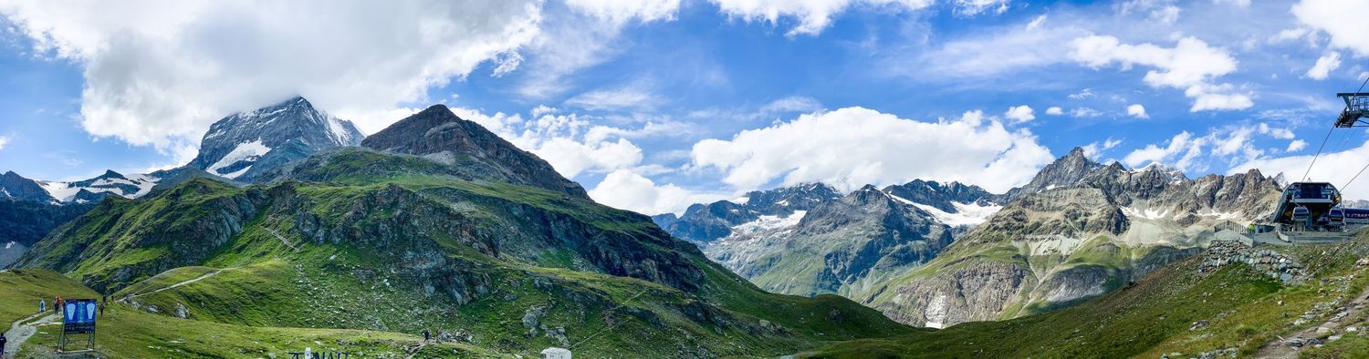 Panoramic mountain vista showing snow-capped peaks and green valleys under blue summer skies.