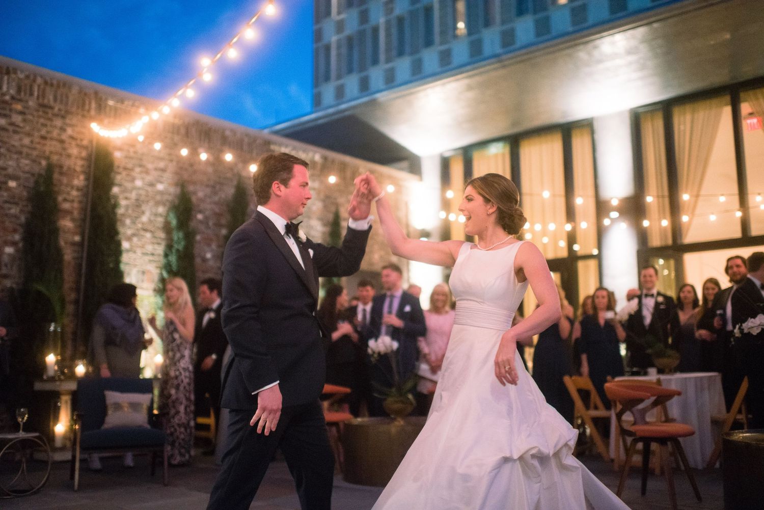 Couple dancing together at an evening outdoor reception with string lights and stone wall backdrop.