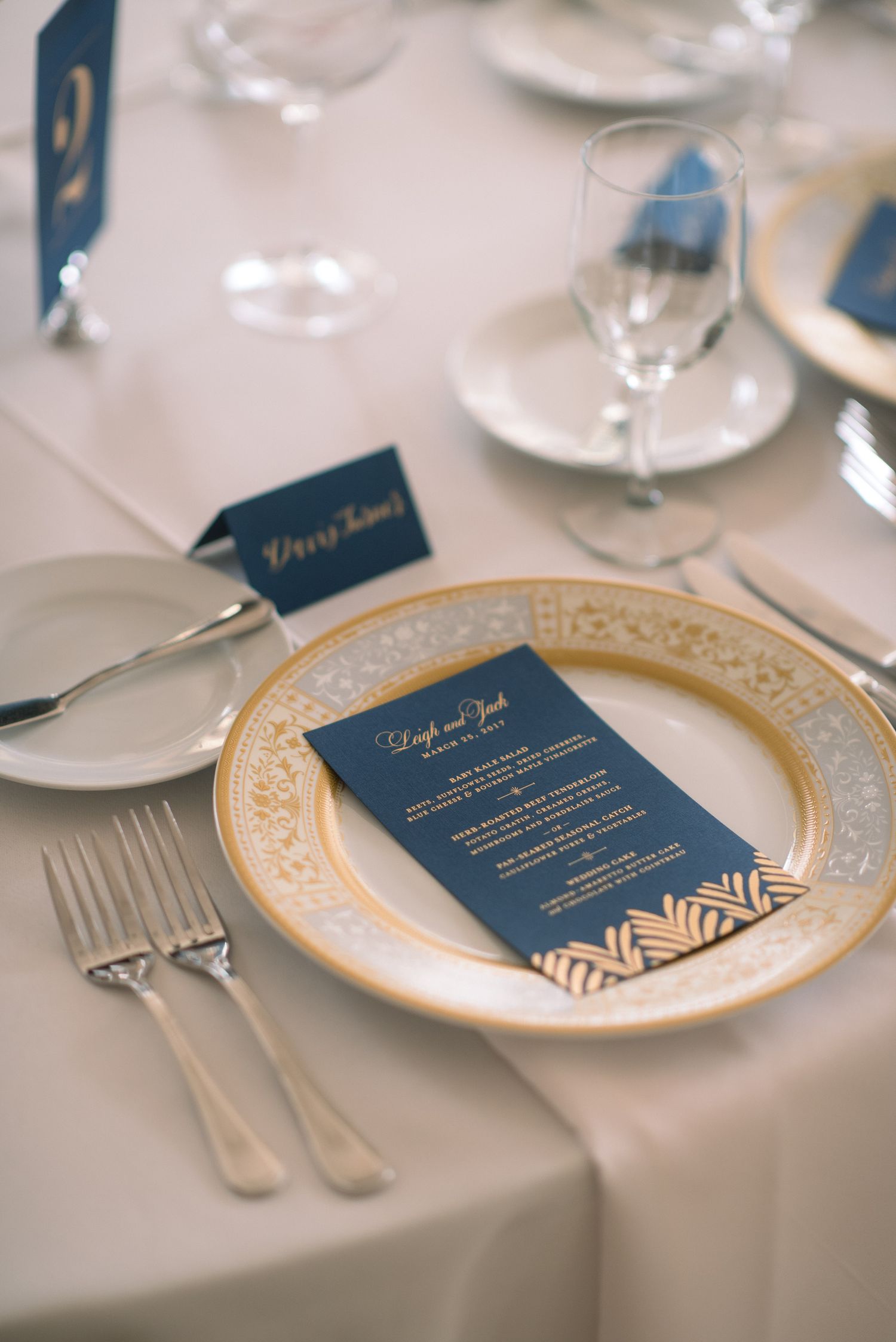 Elegant place settings featuring navy blue menu cards with gold accents on ornate plates at a formal dinner table.