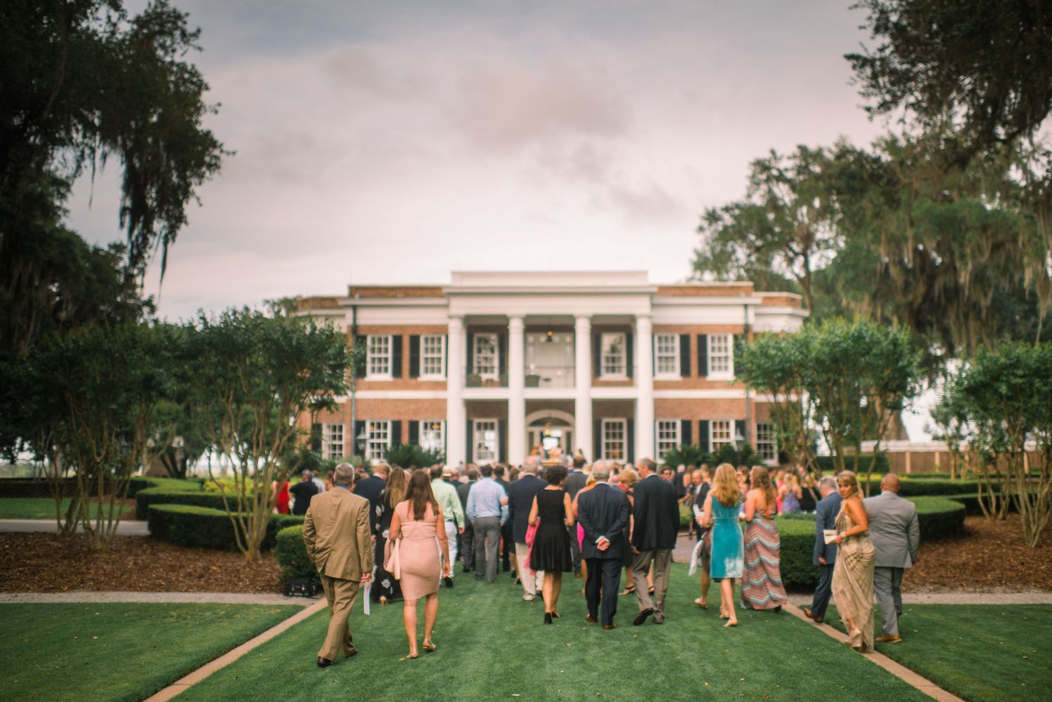 A panoramic view of a stately white columned mansion with wedding guests gathered on the front lawn.