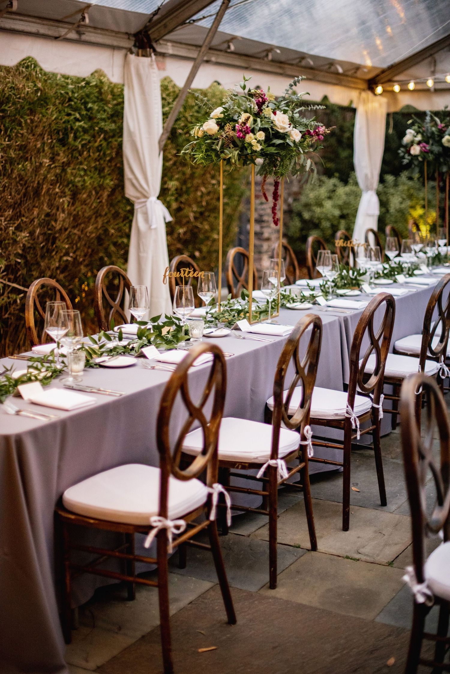Long dining tables with white linens and wooden chairs set up under a covered outdoor wedding reception area.