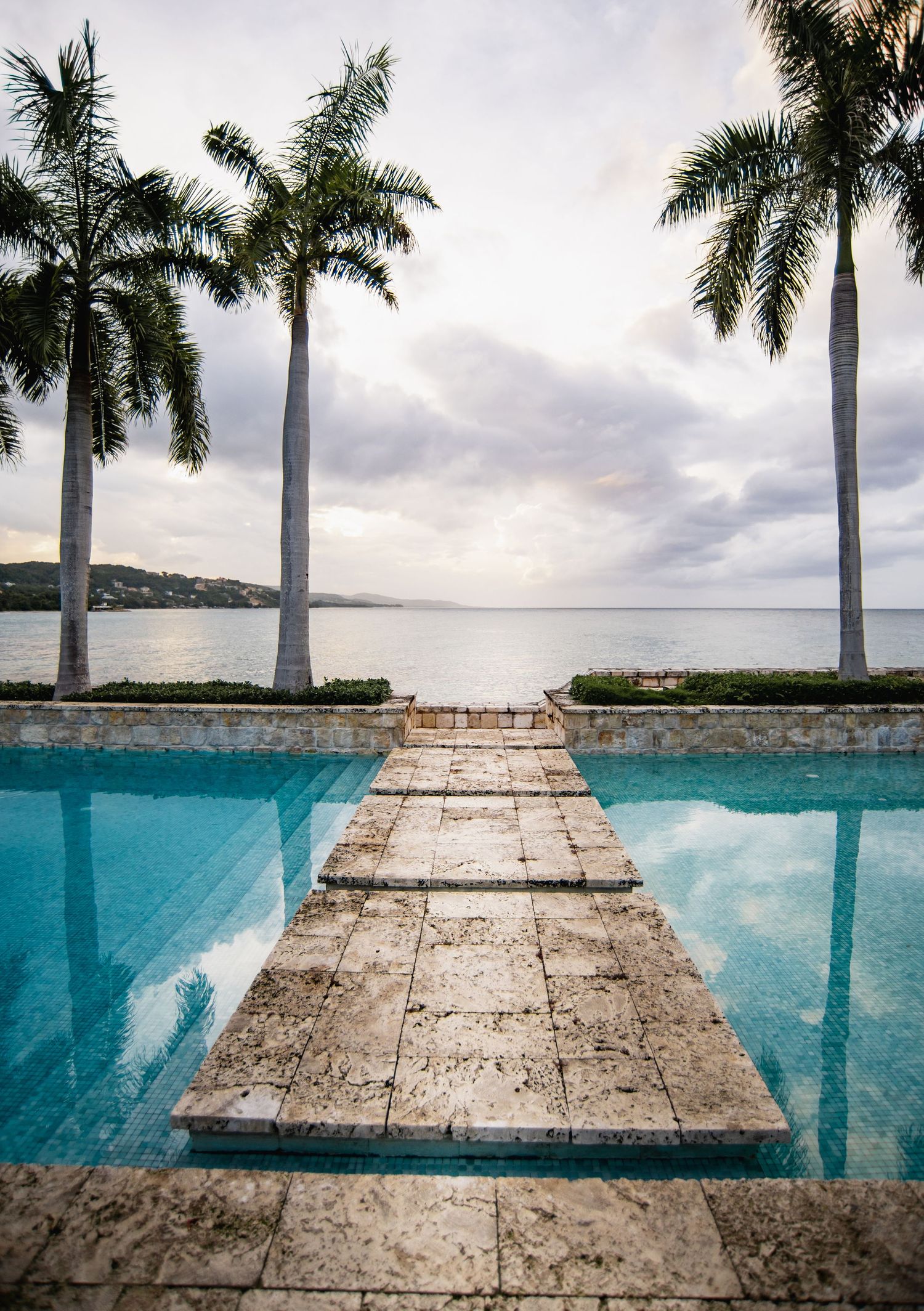 Infinity pool with stone walkway leading to ocean view framed by tropical palm trees under cloudy sky.