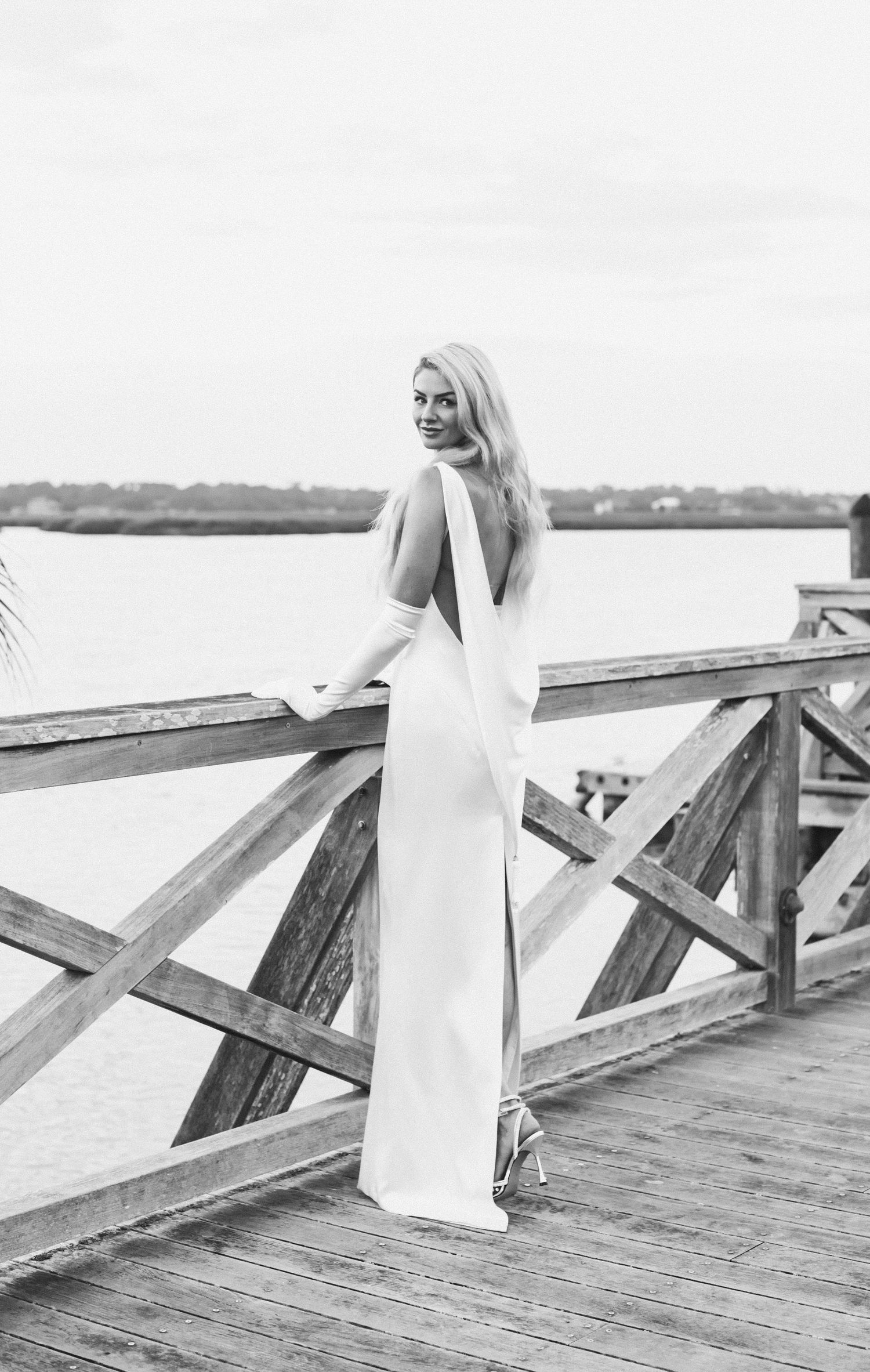 A person in a flowing white dress walks along a rustic wooden pier in black and white photography.