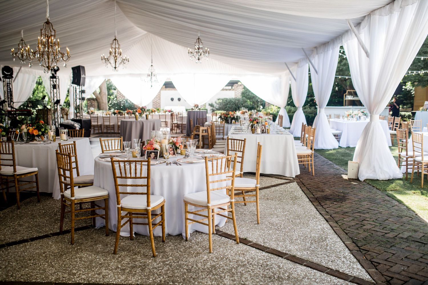 Elegant wedding reception tent setup with white draping, chandeliers, and wooden chiavari chairs arranged in rows.