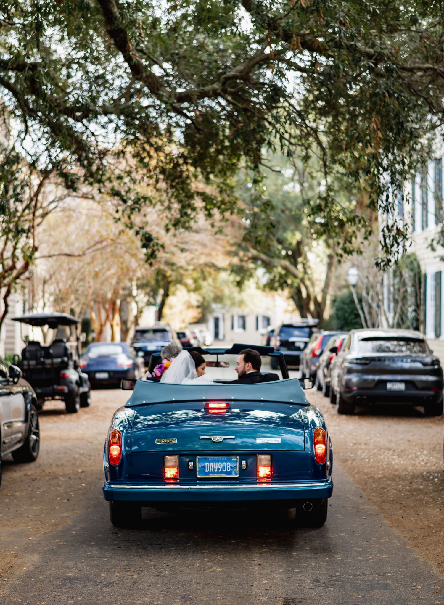 Vintage blue convertible car parked under tree-lined street with other vehicles in background.
