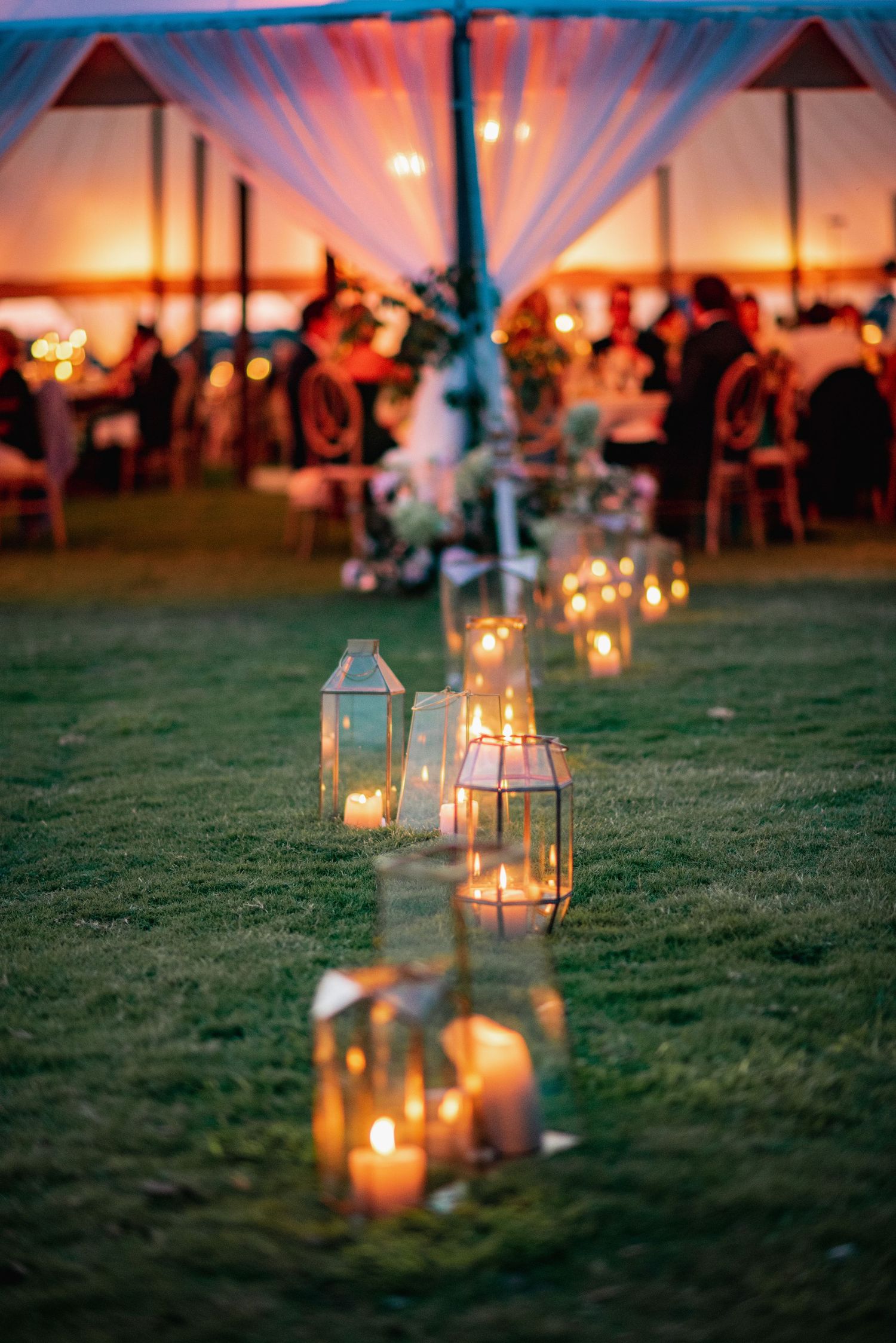 Glowing lanterns line a grassy lawn at dusk with an illuminated party tent in the background creating a romantic ambiance.