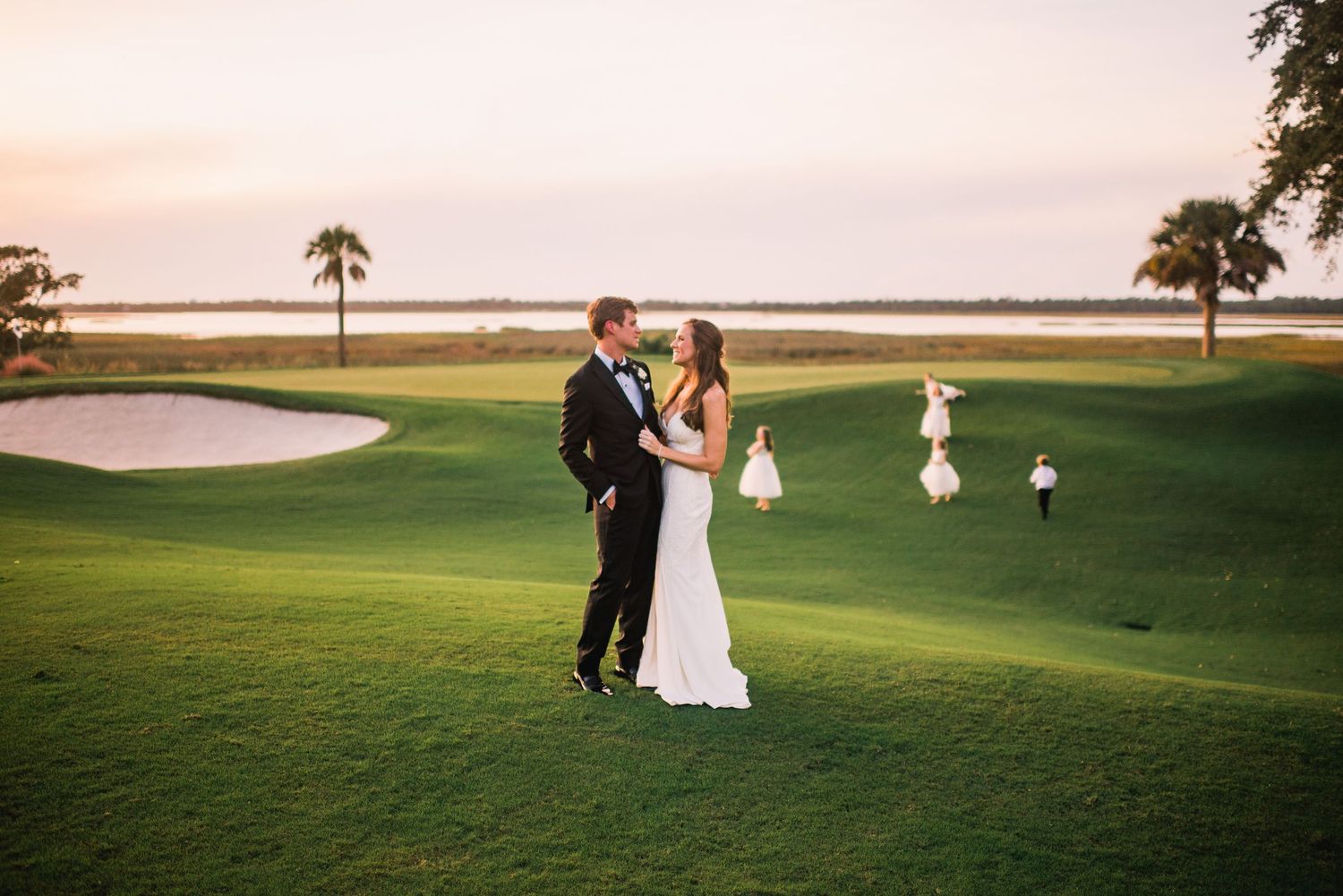 A couple shares a romantic moment on a golf course at sunset with palm trees and water in the background.