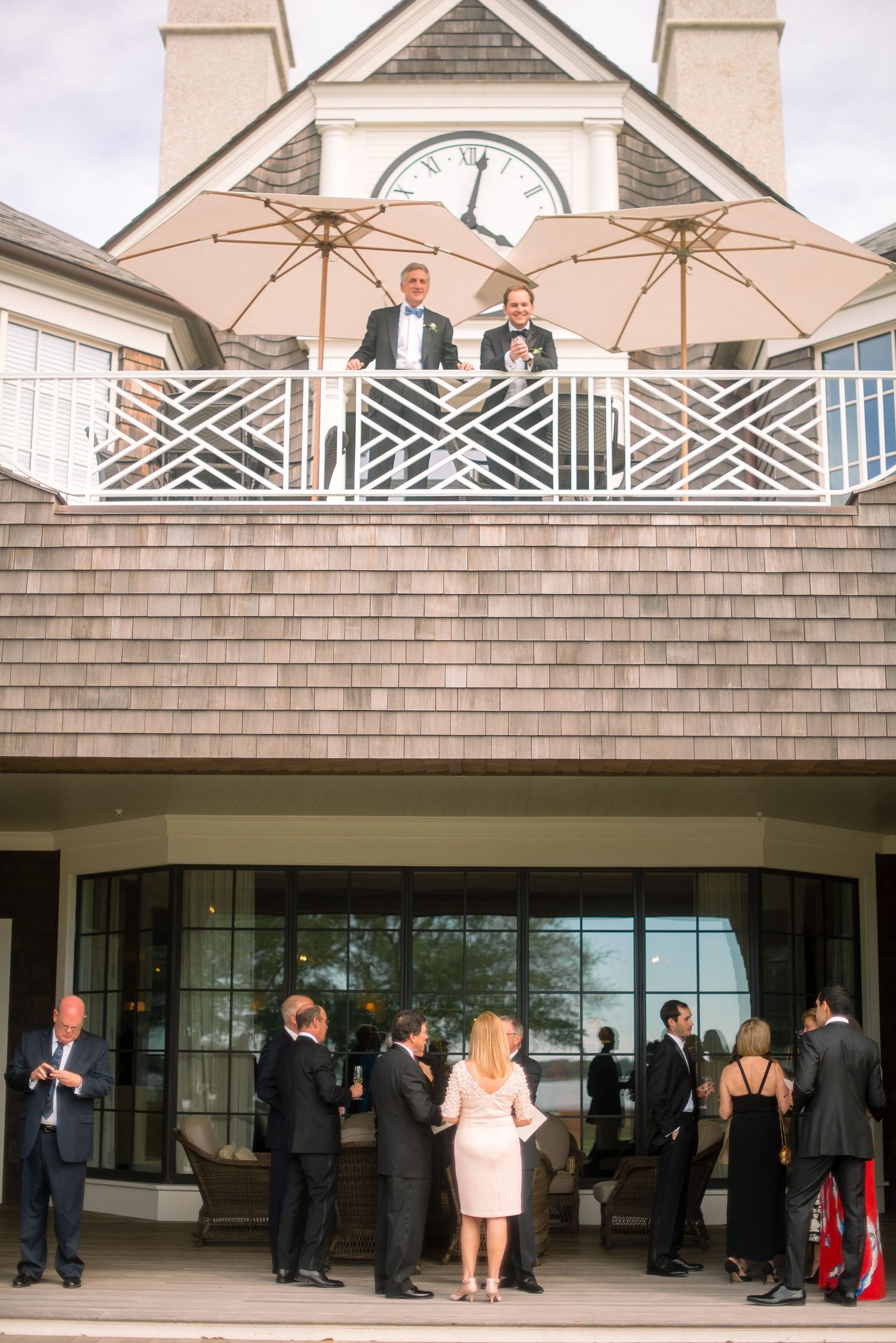 People gather at an elegant coastal venue with two beige umbrellas on an upper balcony and guests mingling below.