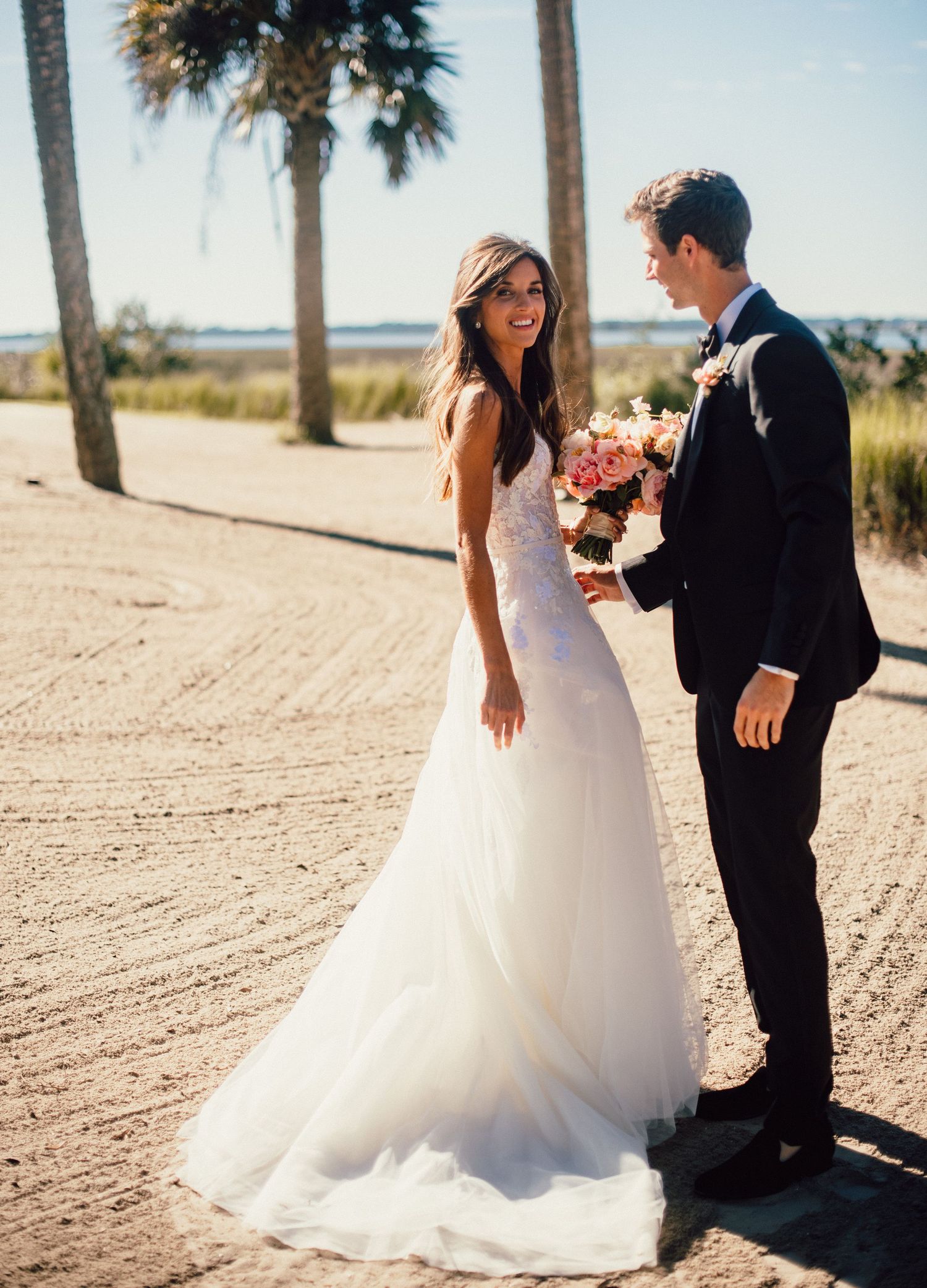 A couple in wedding attire shares a joyful moment together on a sandy beach path lined with palm trees.