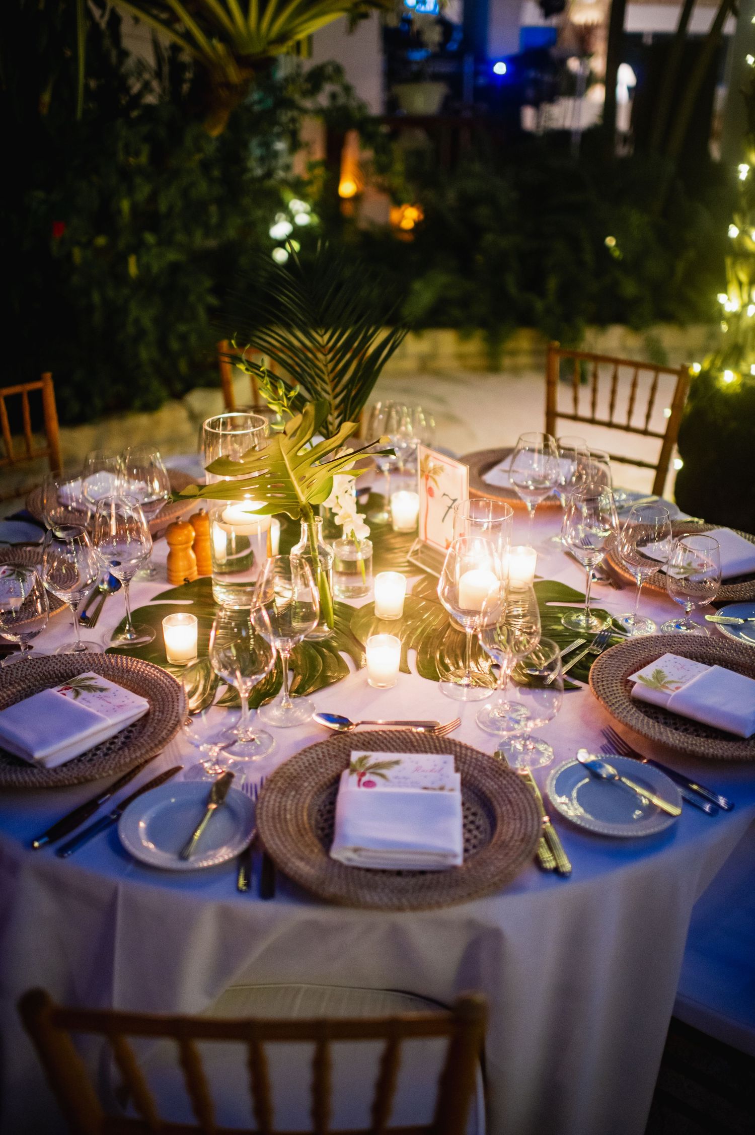 Elegant wedding reception table settings with glowing candles and palm frond decorations at dusk.