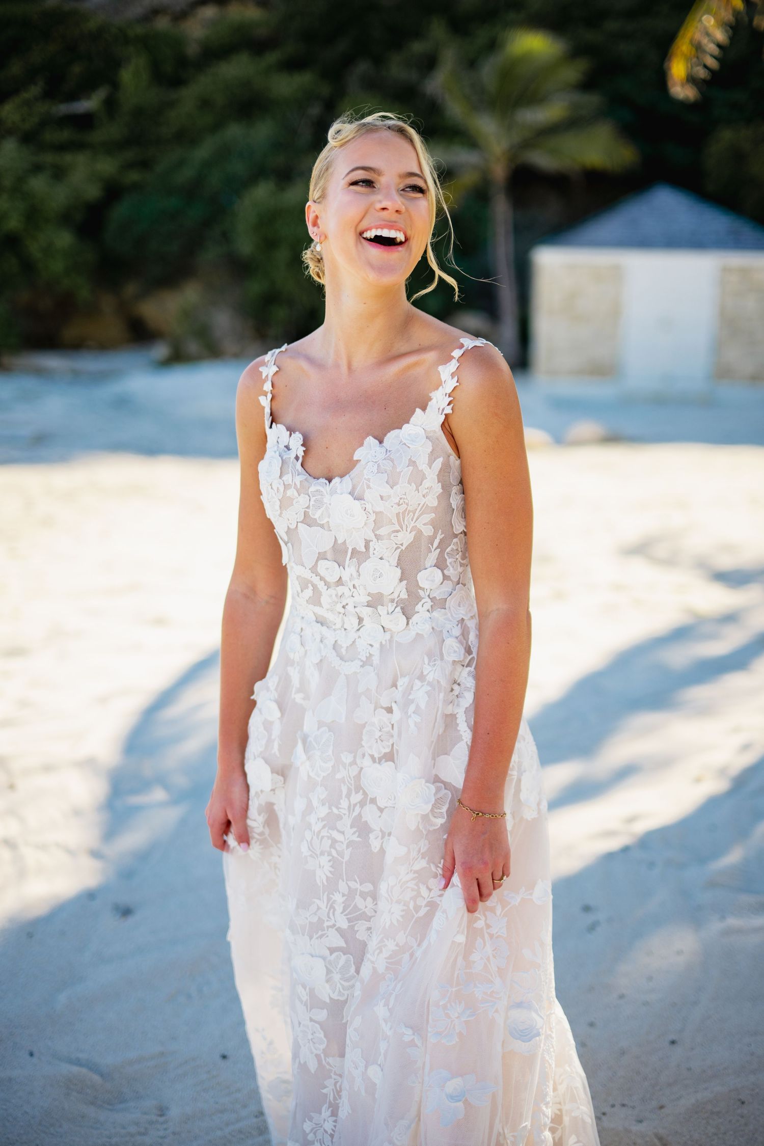 A person in a white lace wedding dress poses joyfully on a sandy beach.