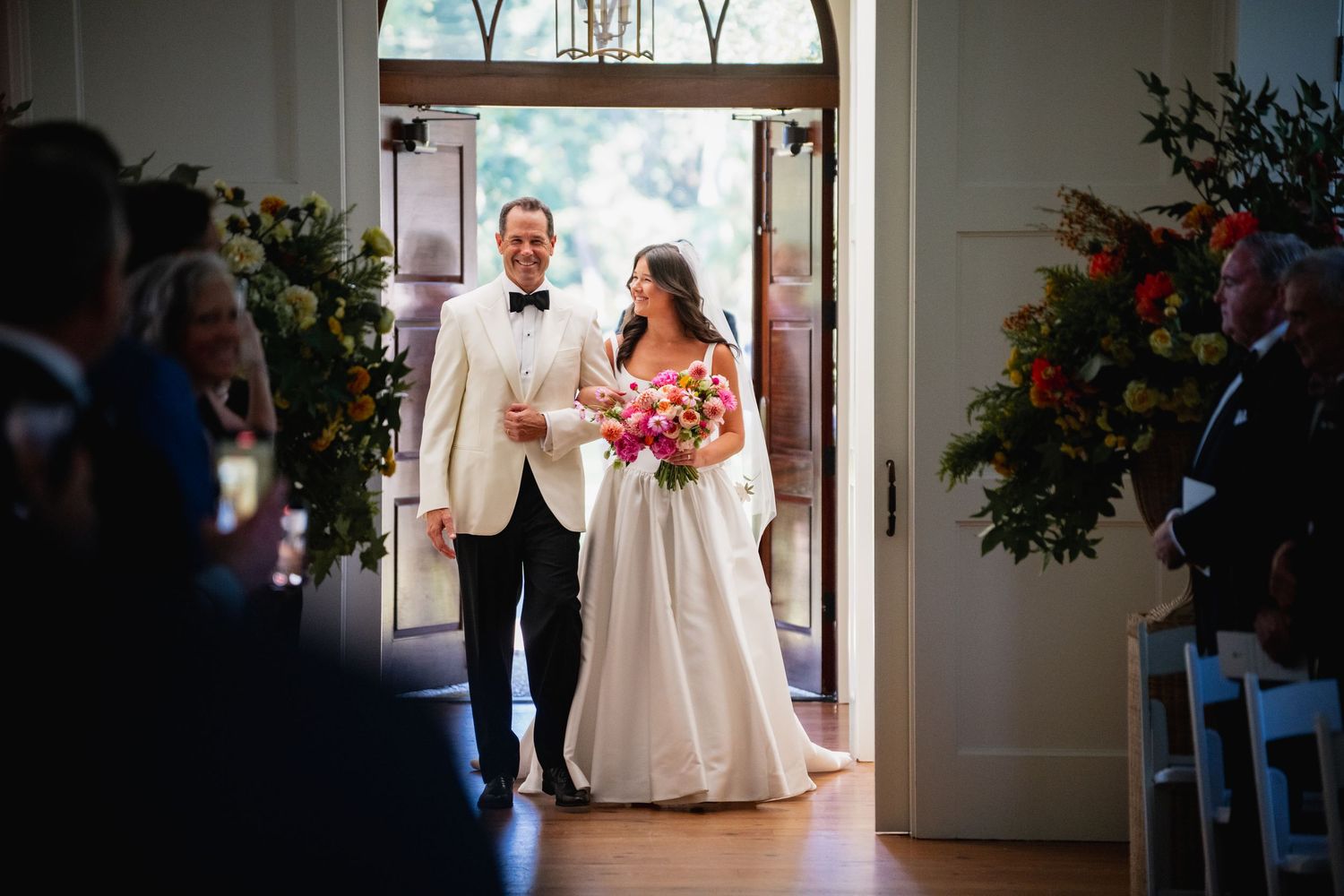 A couple enters through grand wooden doors at their wedding ceremony while holding a vibrant pink bouquet.