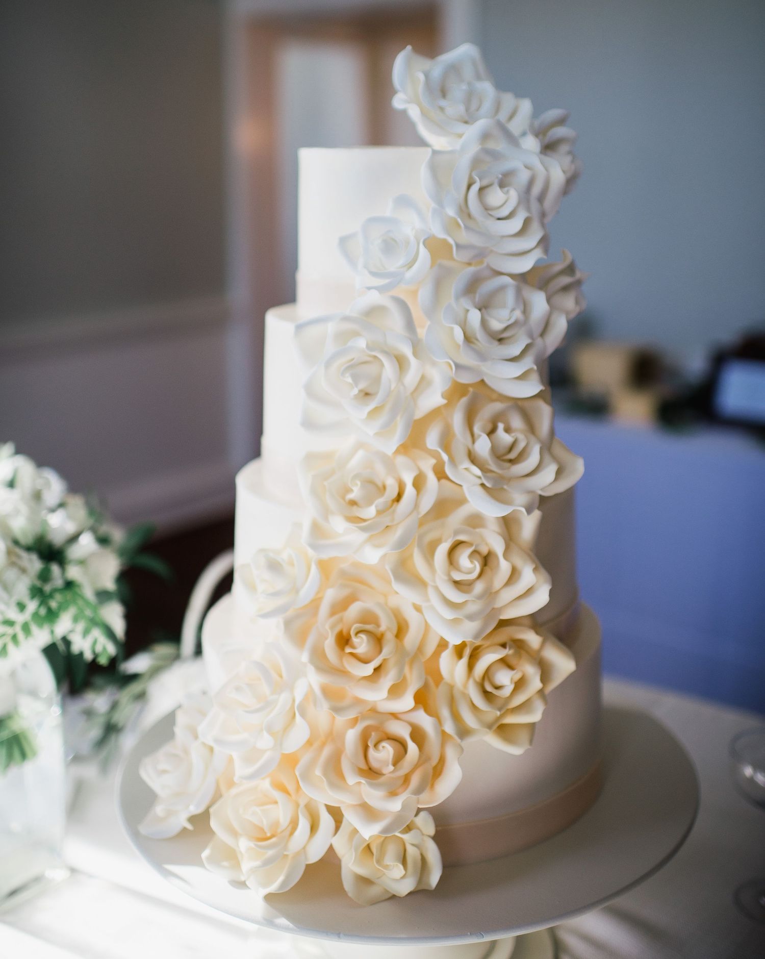 Elegant white wedding cake decorated with cascading sugar roses in ivory and cream tones against a soft blurred background.
