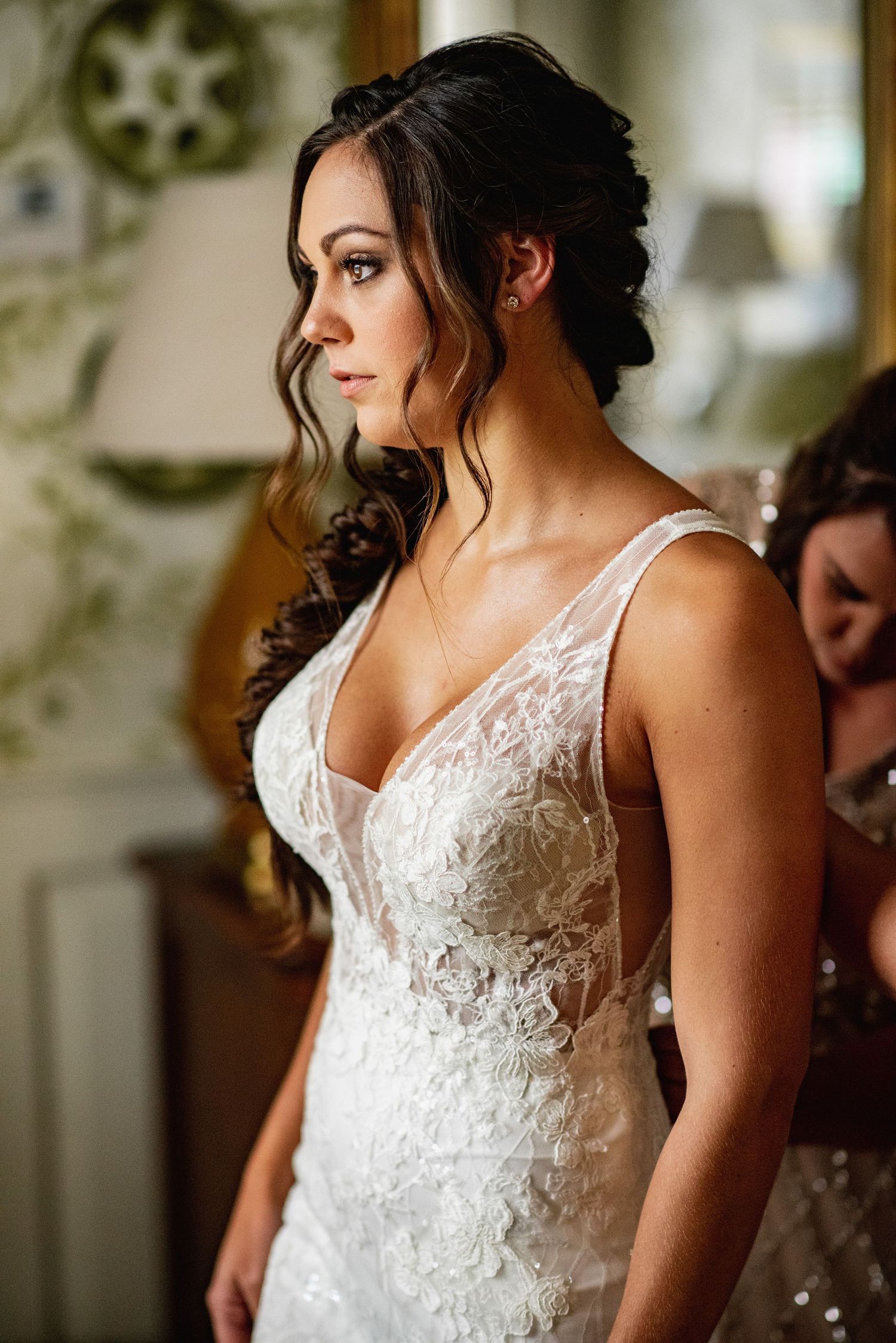 A bride in a white lace wedding dress with delicate straps stands elegantly in soft indoor lighting.