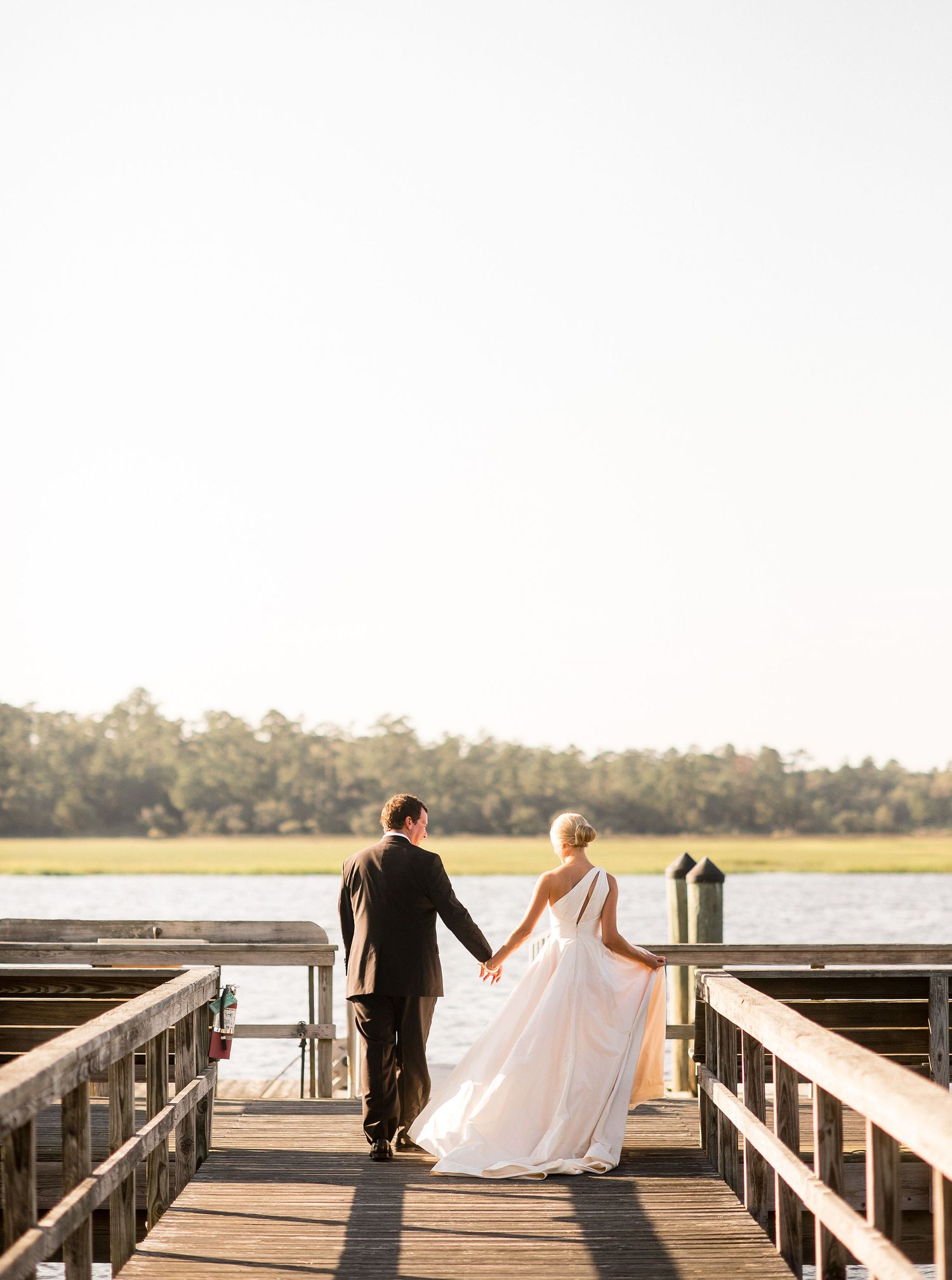 A couple in wedding attire walks hand in hand on a wooden dock overlooking a serene lake at sunset.