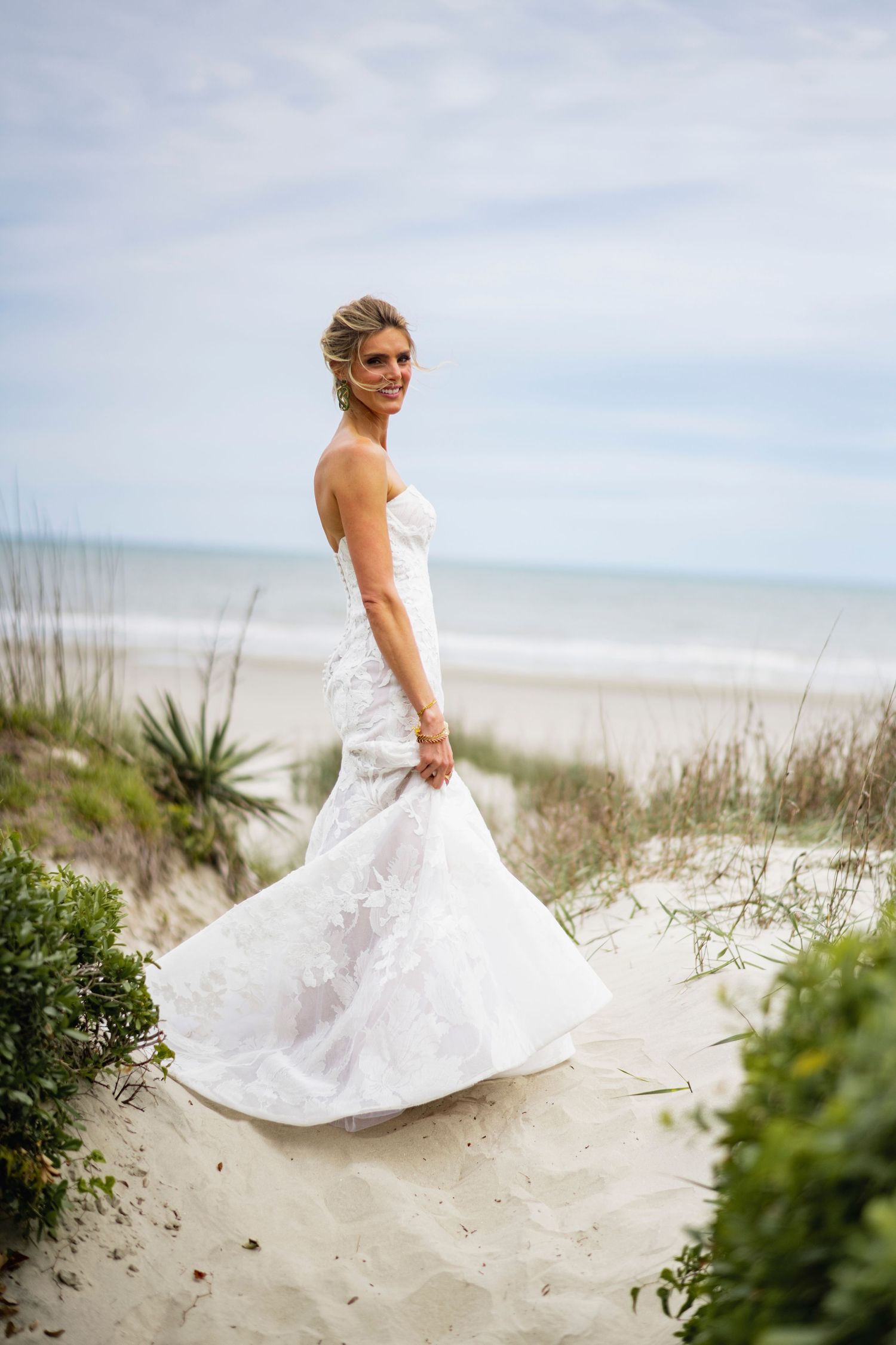 A bride in a white wedding dress twirls gracefully on a sandy beach with sea grass and ocean waves in the background.