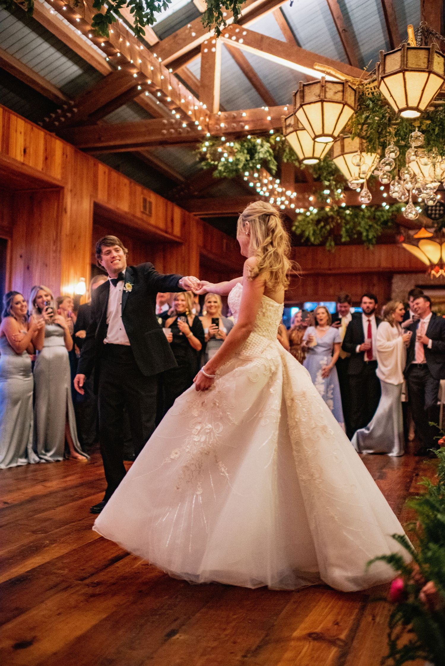 Newlyweds share their first dance in a rustic barn venue decorated with twinkling lights and greenery.