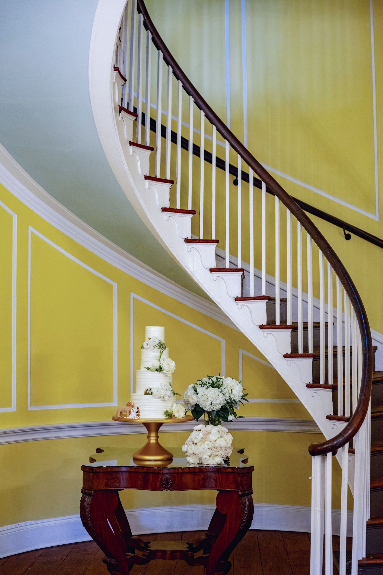 Elegant curved staircase with dark wood banister against yellow walls, featuring antique table with wedding cake and flowers.