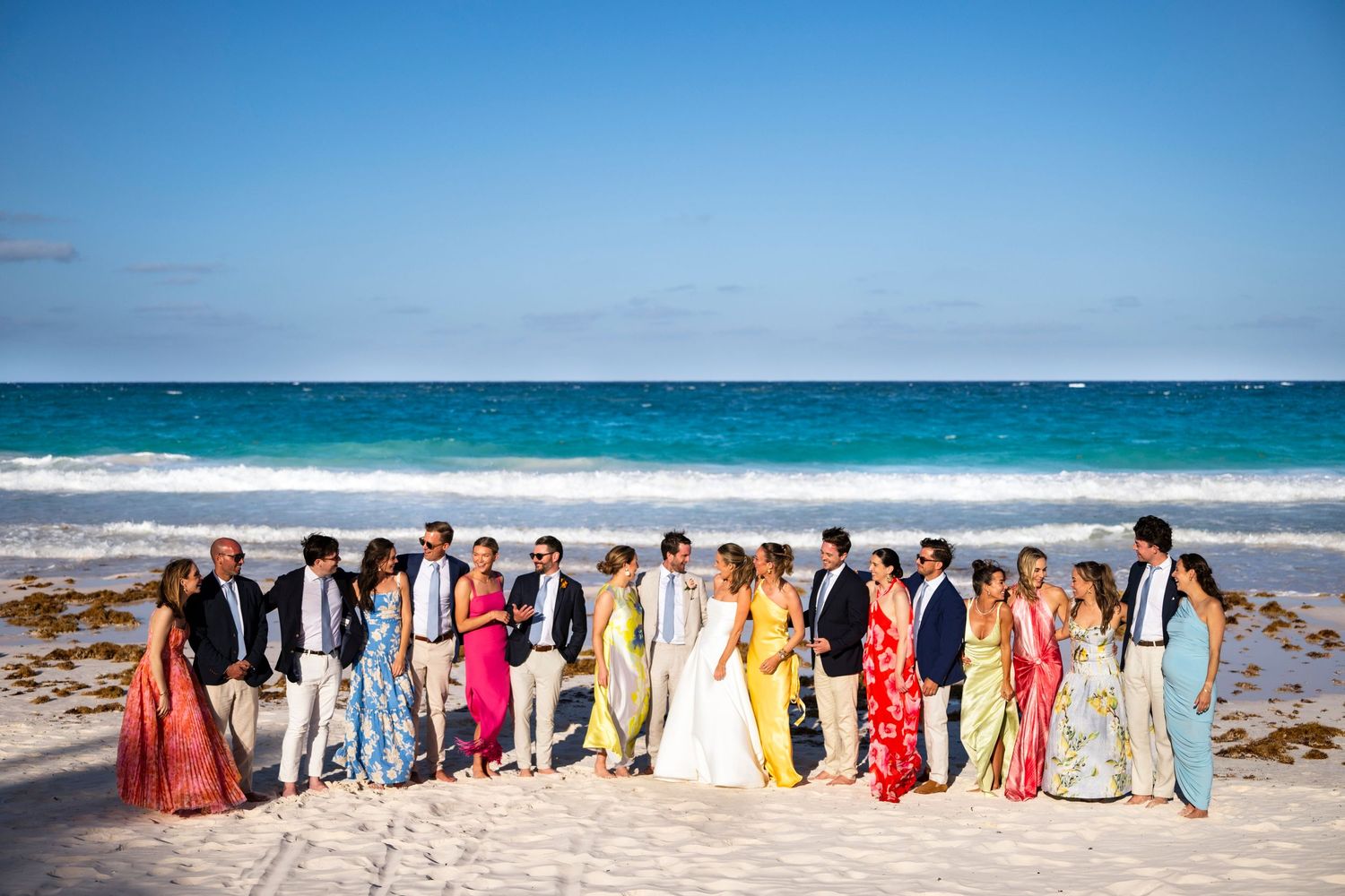A panoramic sequence of photos showing a large group of people in colorful formal attire standing on a sunny beach.