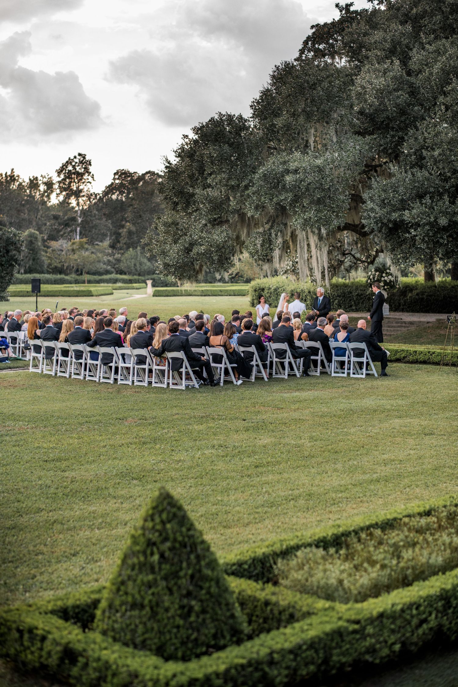 Outdoor wedding ceremony setup with white chairs arranged in rows on a lush green lawn surrounded by mature trees.