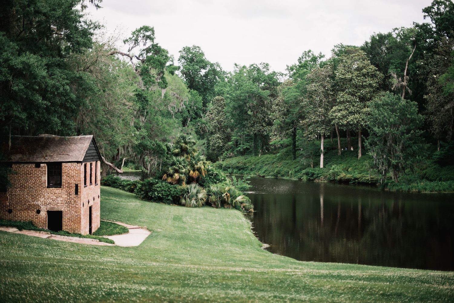 Small brick structure sits on manicured lawn beside tranquil pond surrounded by lush green forest.