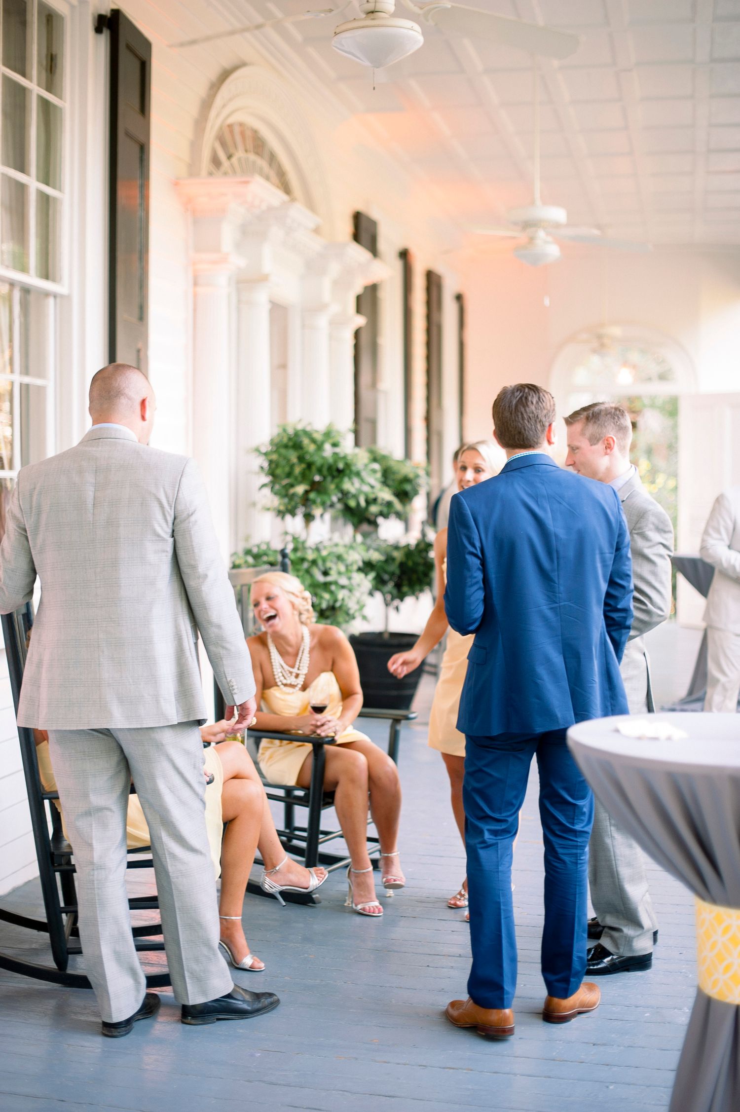 Wedding guests mingle and socialize in an elegant columned hallway with soft pink lighting and white architecture.