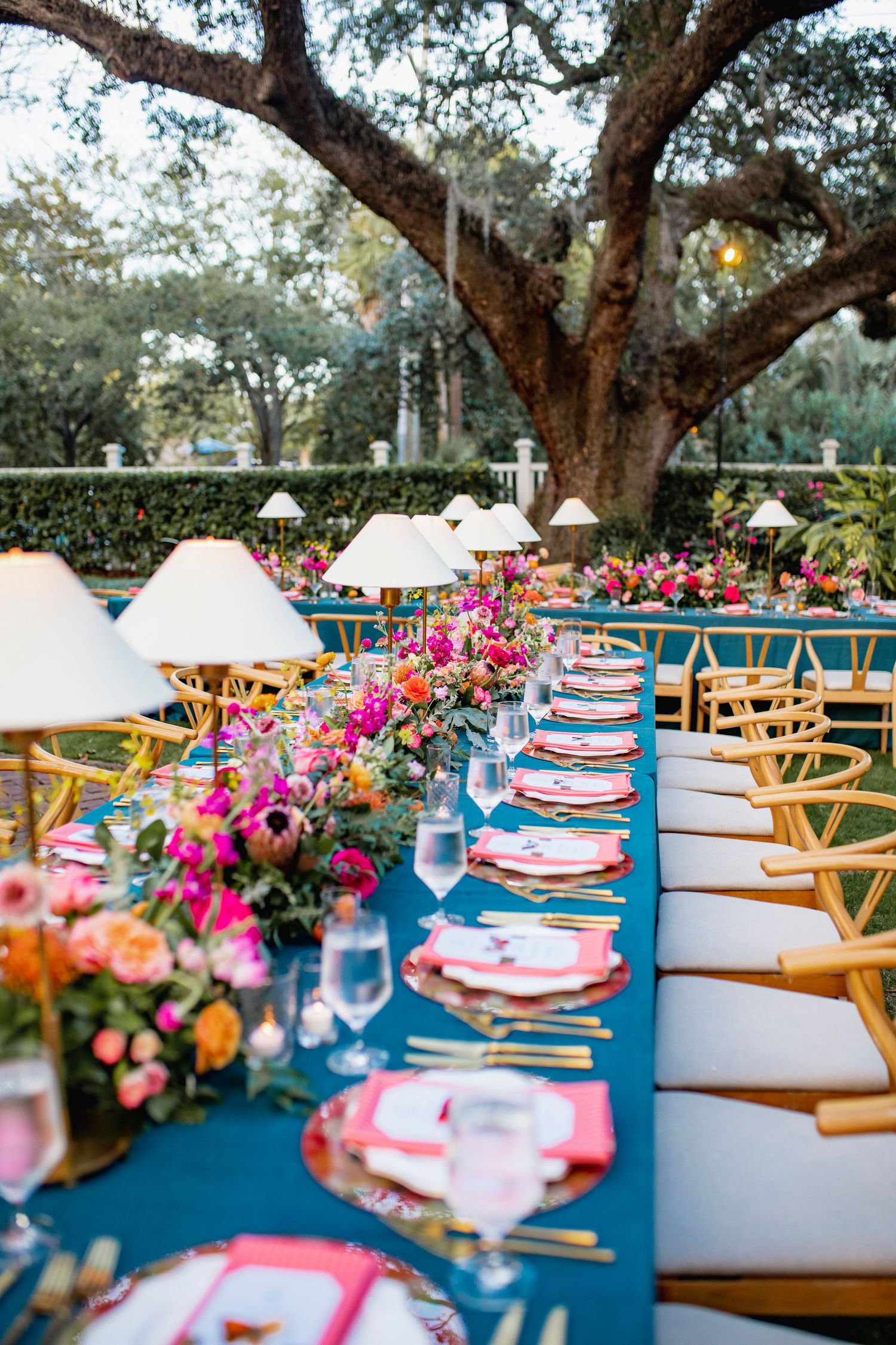 Elegant outdoor wedding reception table with navy blue linens, bright pink floral centerpieces and wooden chairs under oak trees.