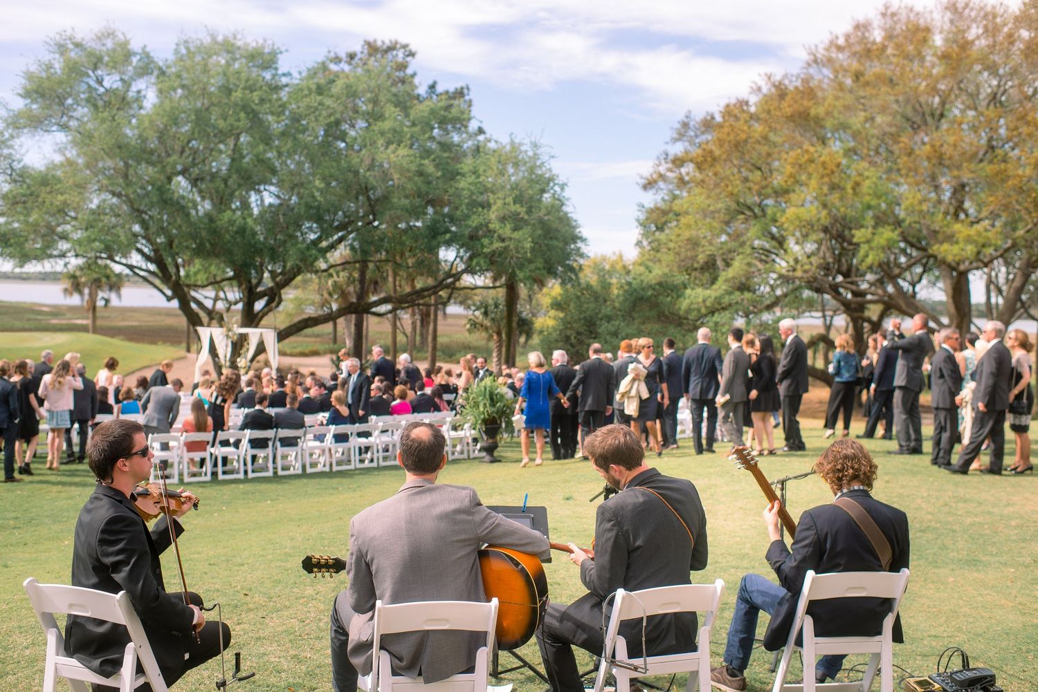 Musicians perform at an outdoor gathering with a large crowd of people enjoying music in a park setting under autumn trees.