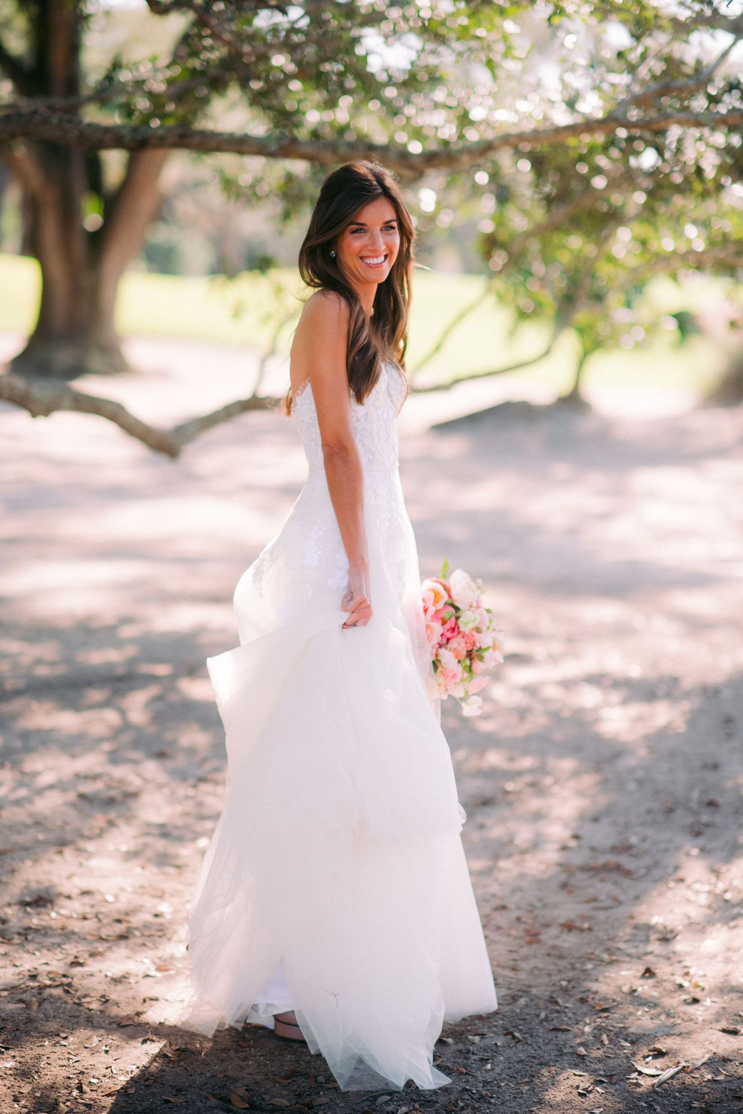 A bride in a flowing white wedding dress poses outdoors under blossoming trees in a romantic garden setting.