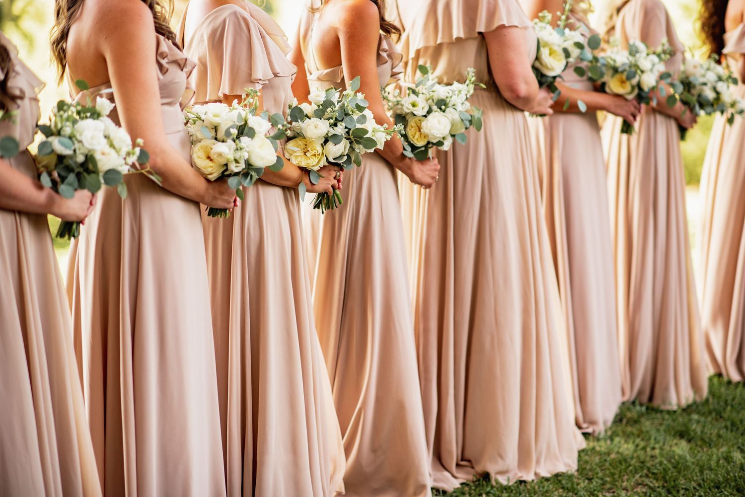 Bridesmaids in blush pink dresses stand in a row holding white floral bouquets during an outdoor wedding ceremony.