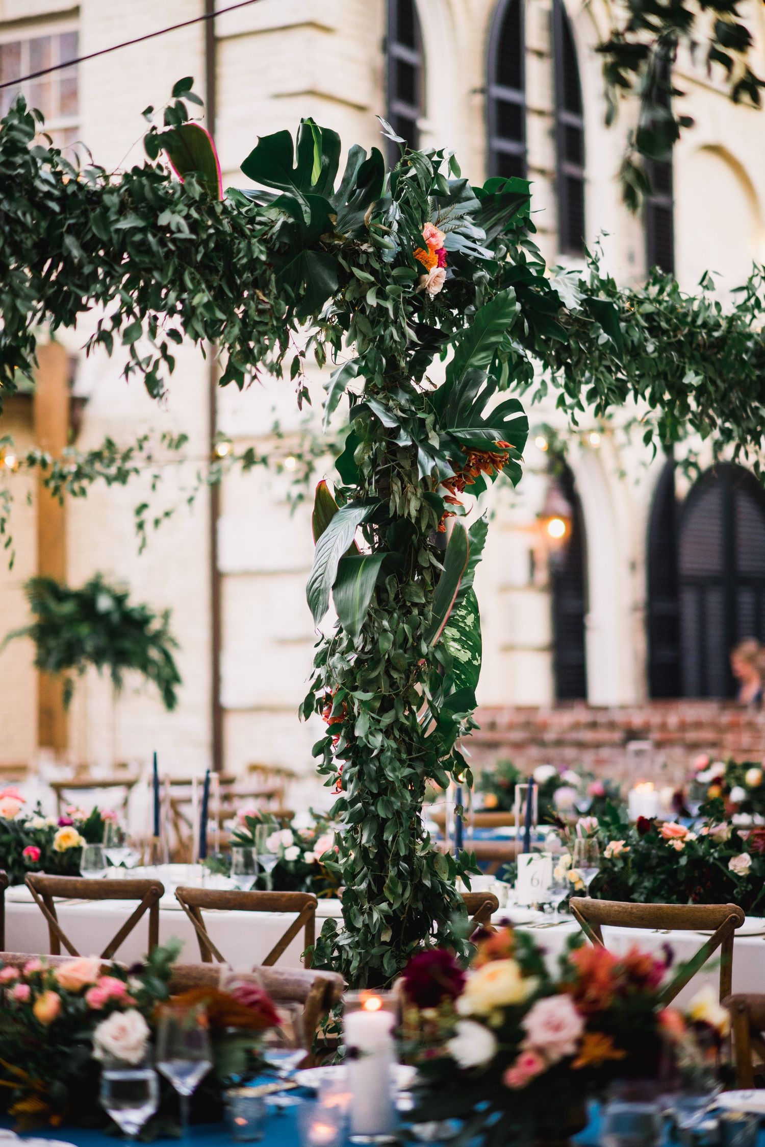 Elegant wedding reception table with lush greenery arch, blue tablecloth, and floral centerpieces in an outdoor venue.