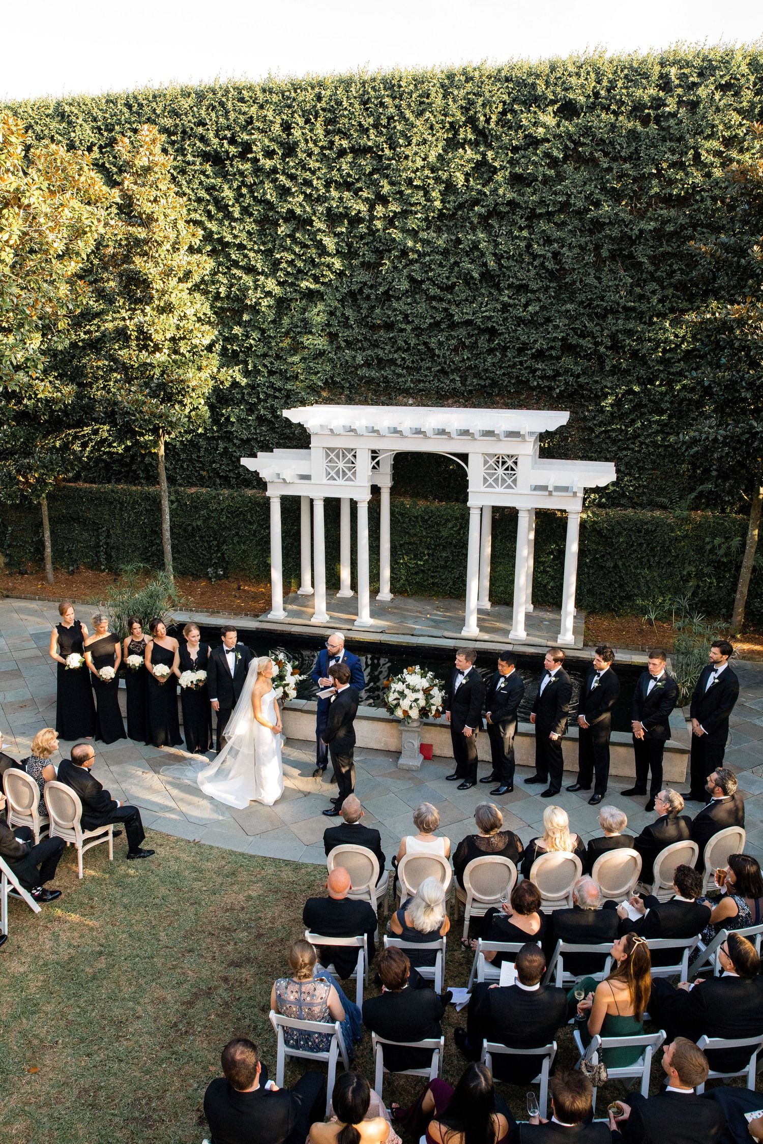 An elegant outdoor wedding ceremony setup with white columns and chairs arranged on a lawn surrounded by hedges.