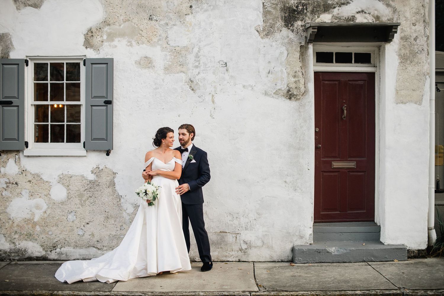 A bride and groom share an intimate moment against a weathered white stone wall with rustic shuttered windows.