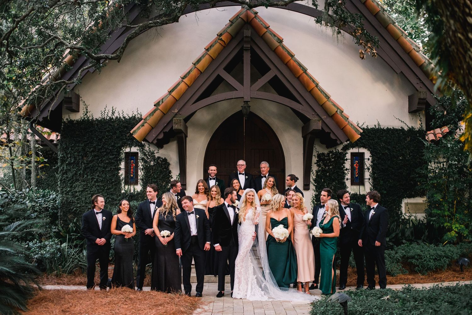 Wedding party poses outdoors in formal attire in front of a chapel with gothic architecture and greenery.