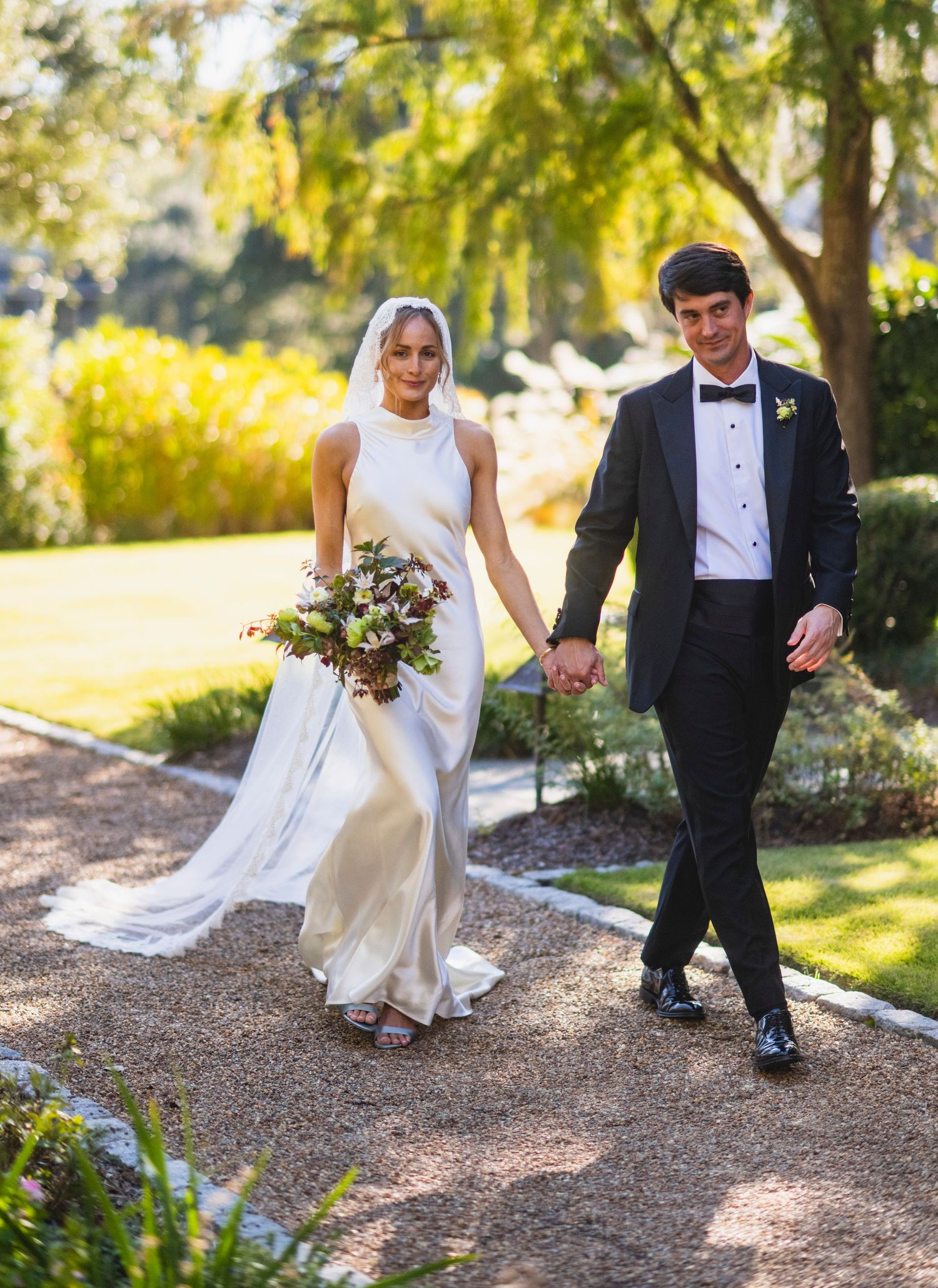 A couple in wedding attire playfully walking and dancing together on a gravel path surrounded by autumn trees.