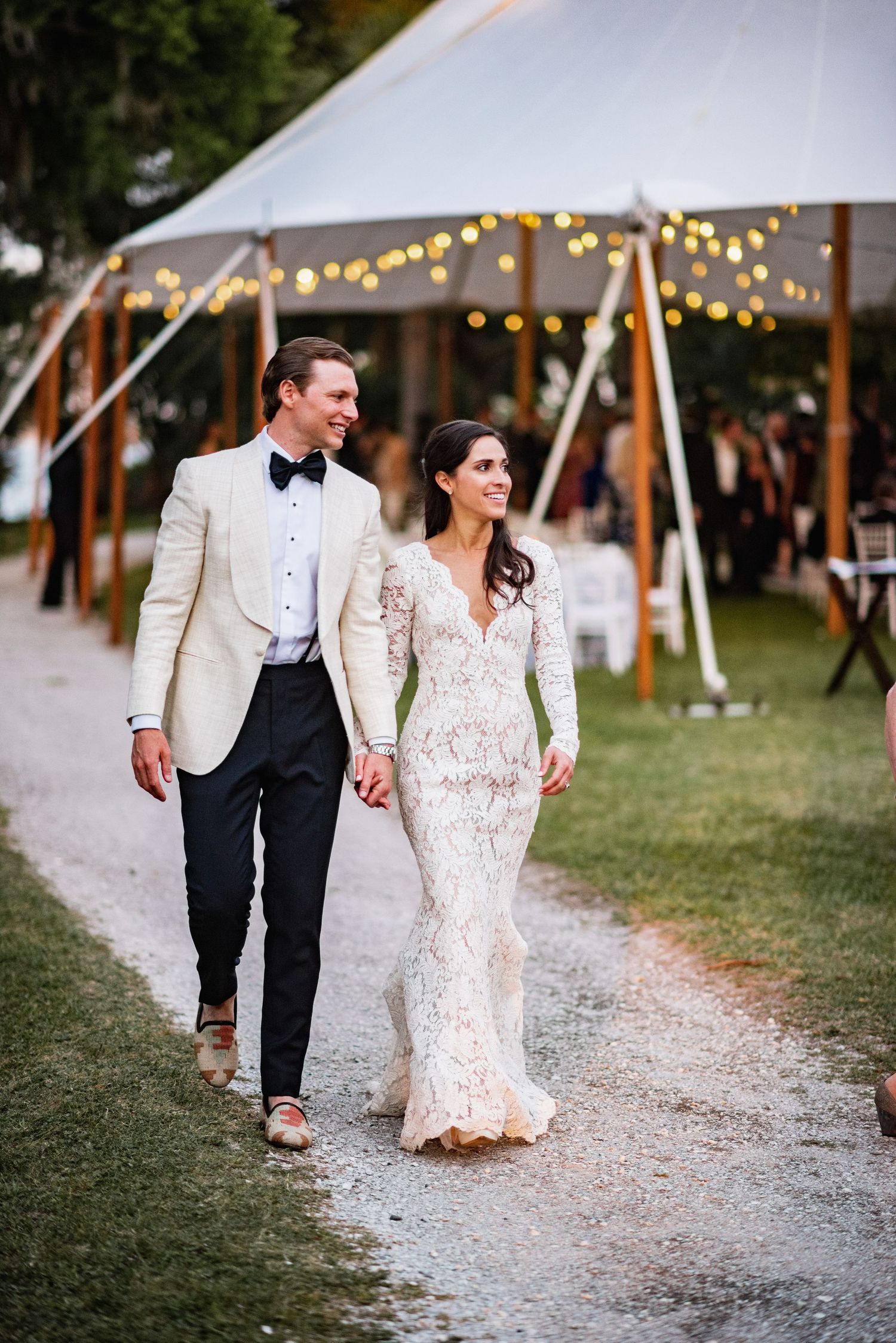 Newlywed couple walking hand in hand on gravel path under string lights at outdoor wedding reception.