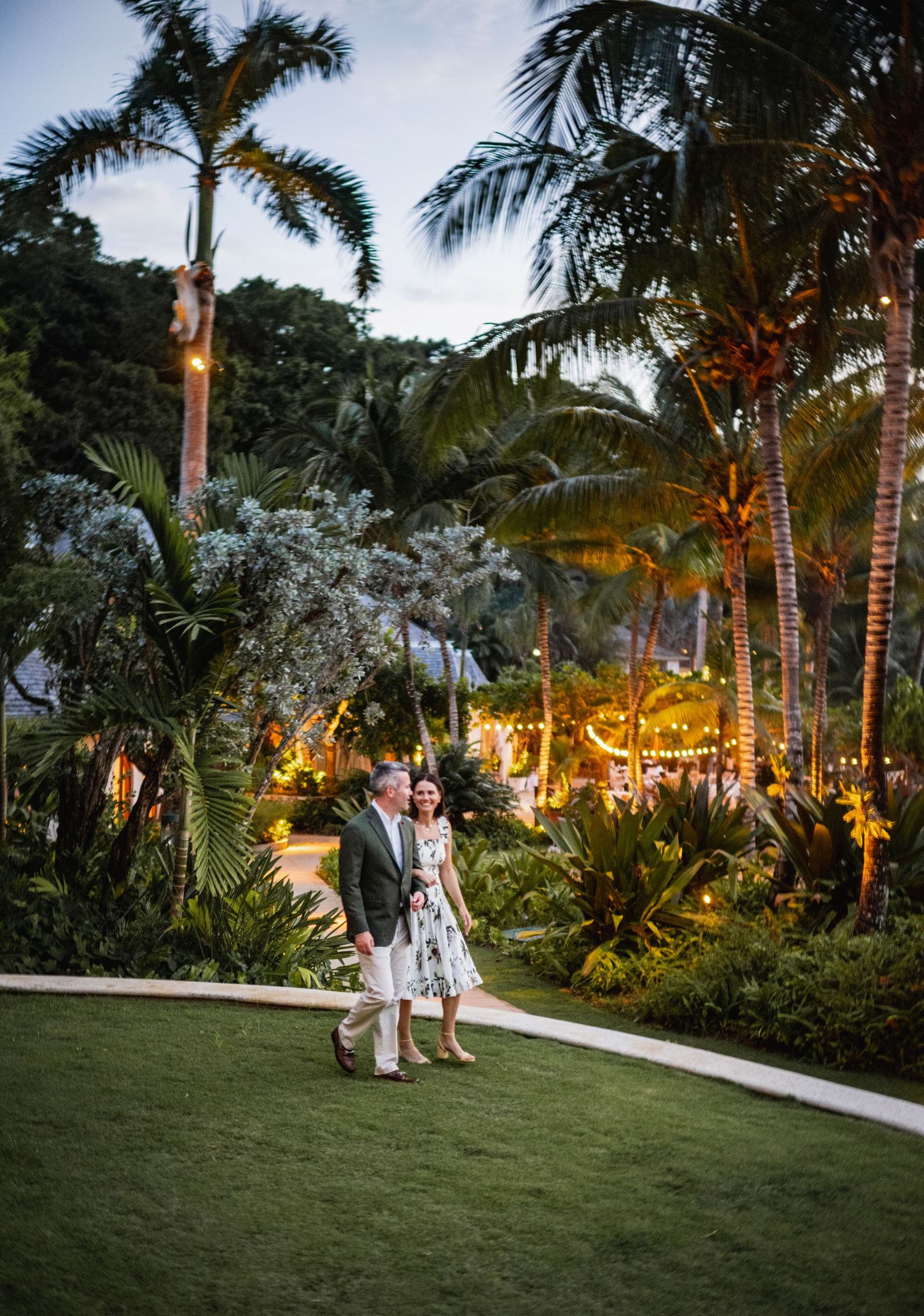 Romantic evening garden party setting with palm trees illuminated by warm lights and couples enjoying the tropical atmosphere.