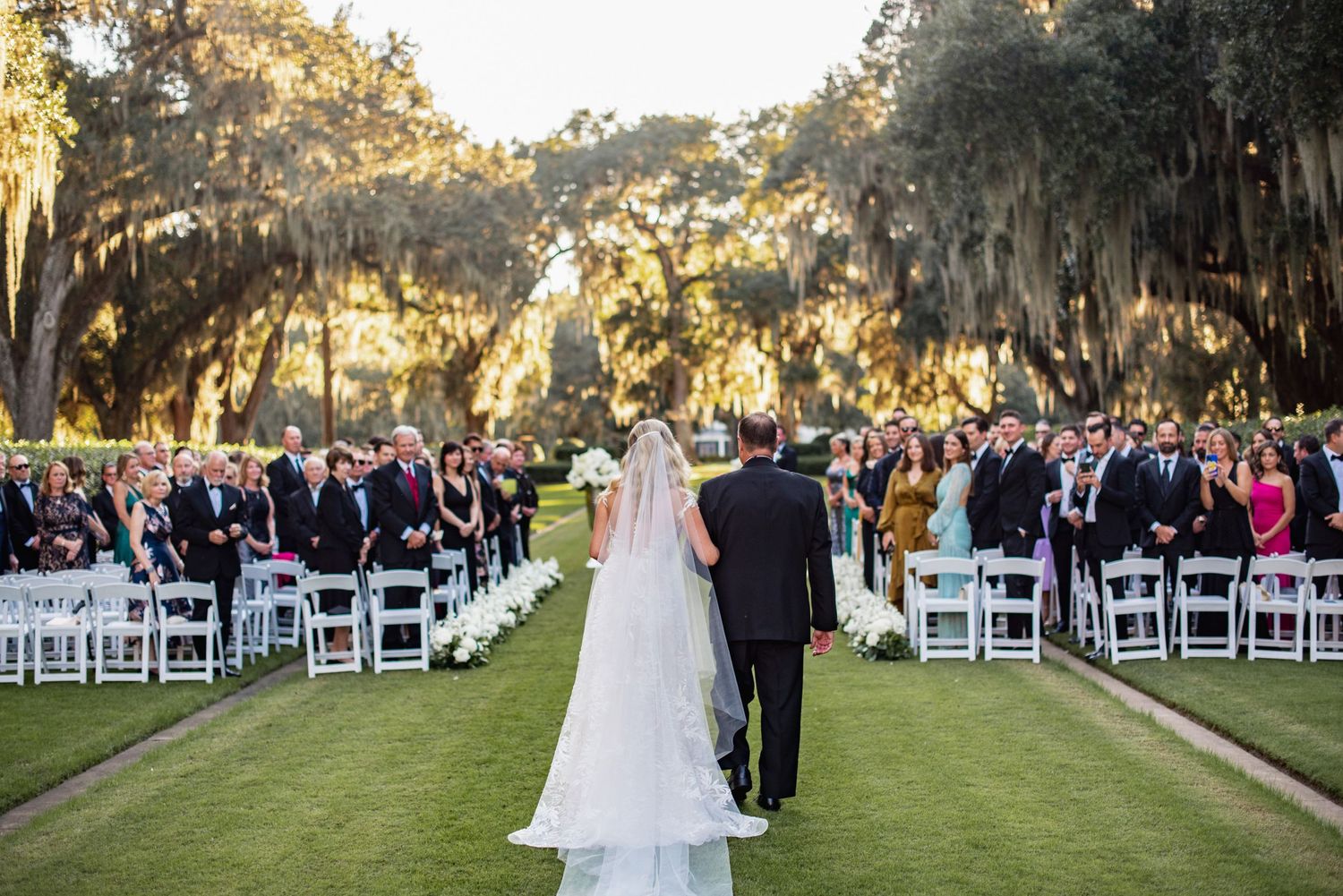 Outdoor wedding ceremony setup with white chairs arranged in rows under Spanish moss-draped oak trees at sunset.