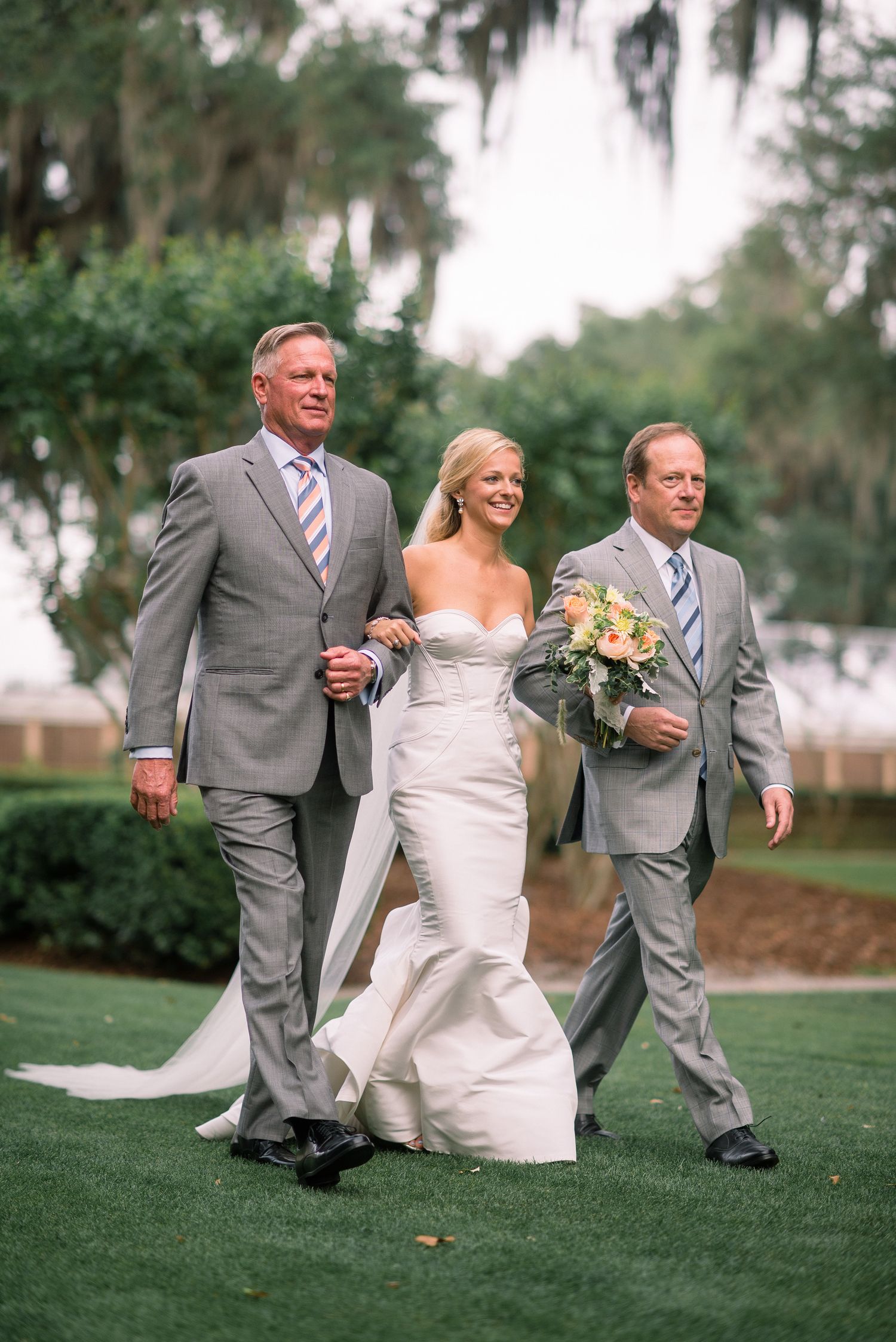 Wedding party in gray suits and white dress walking across a grassy lawn at an outdoor ceremony.