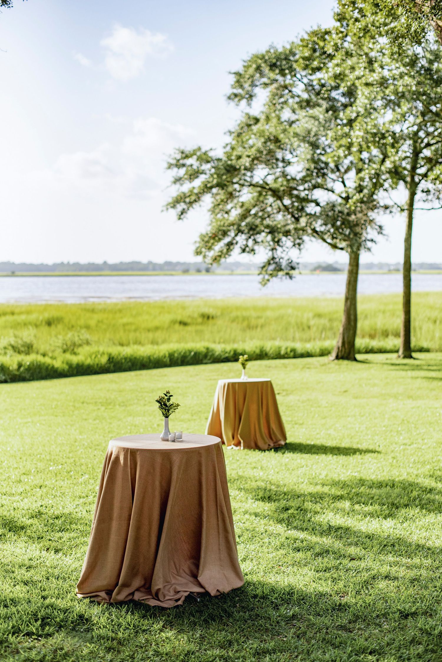 Elegant round tables with gold tablecloths set up on a waterfront lawn for an outdoor event.