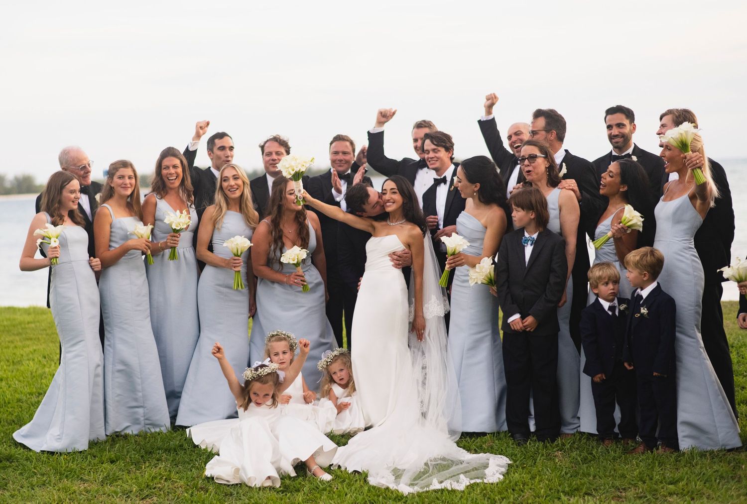 Wedding party in formal attire poses joyfully on a grassy lawn by the water during an outdoor celebration.