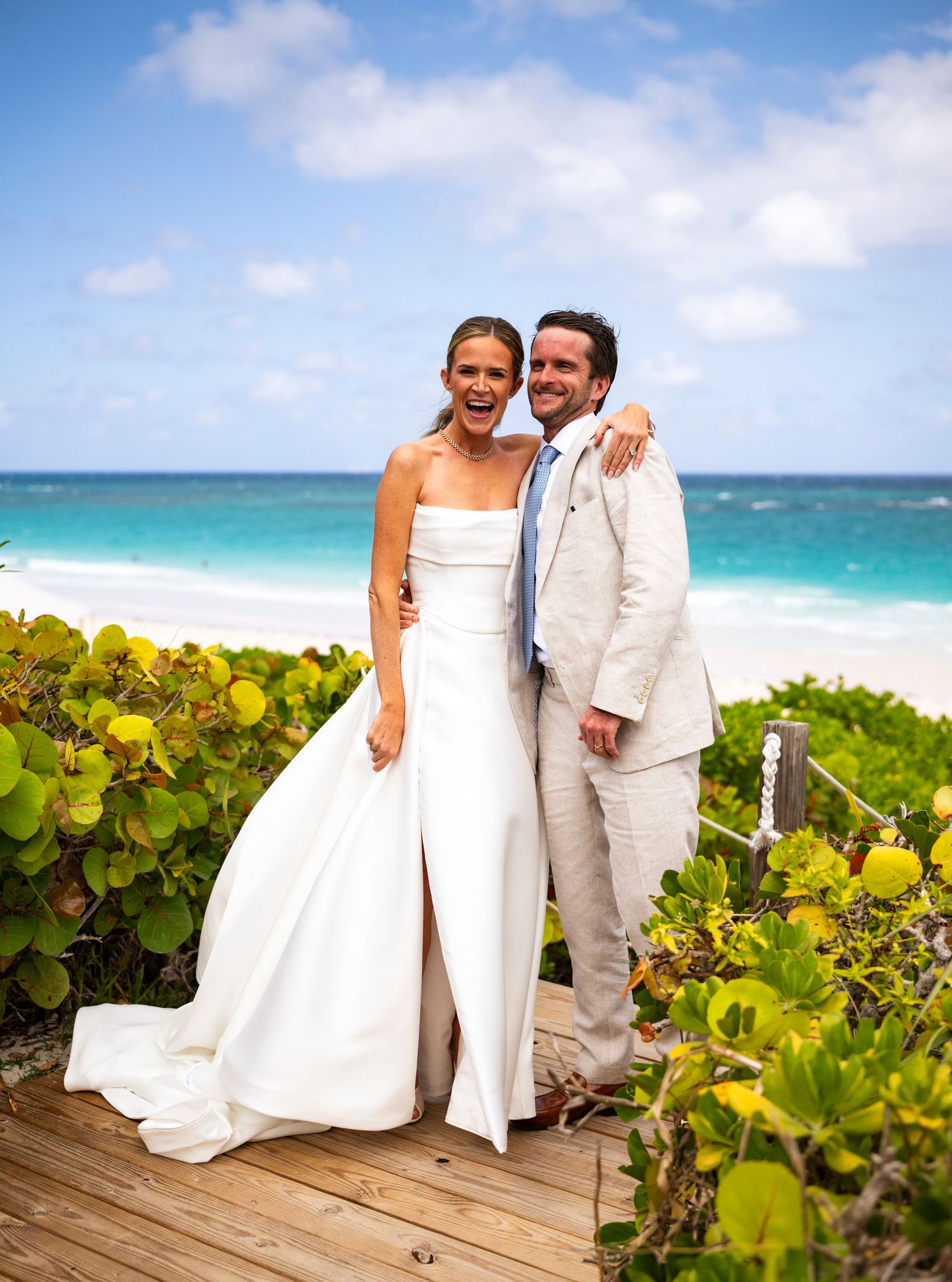 A couple in wedding attire poses along a beach boardwalk with lush sea grape plants and turquoise ocean views.