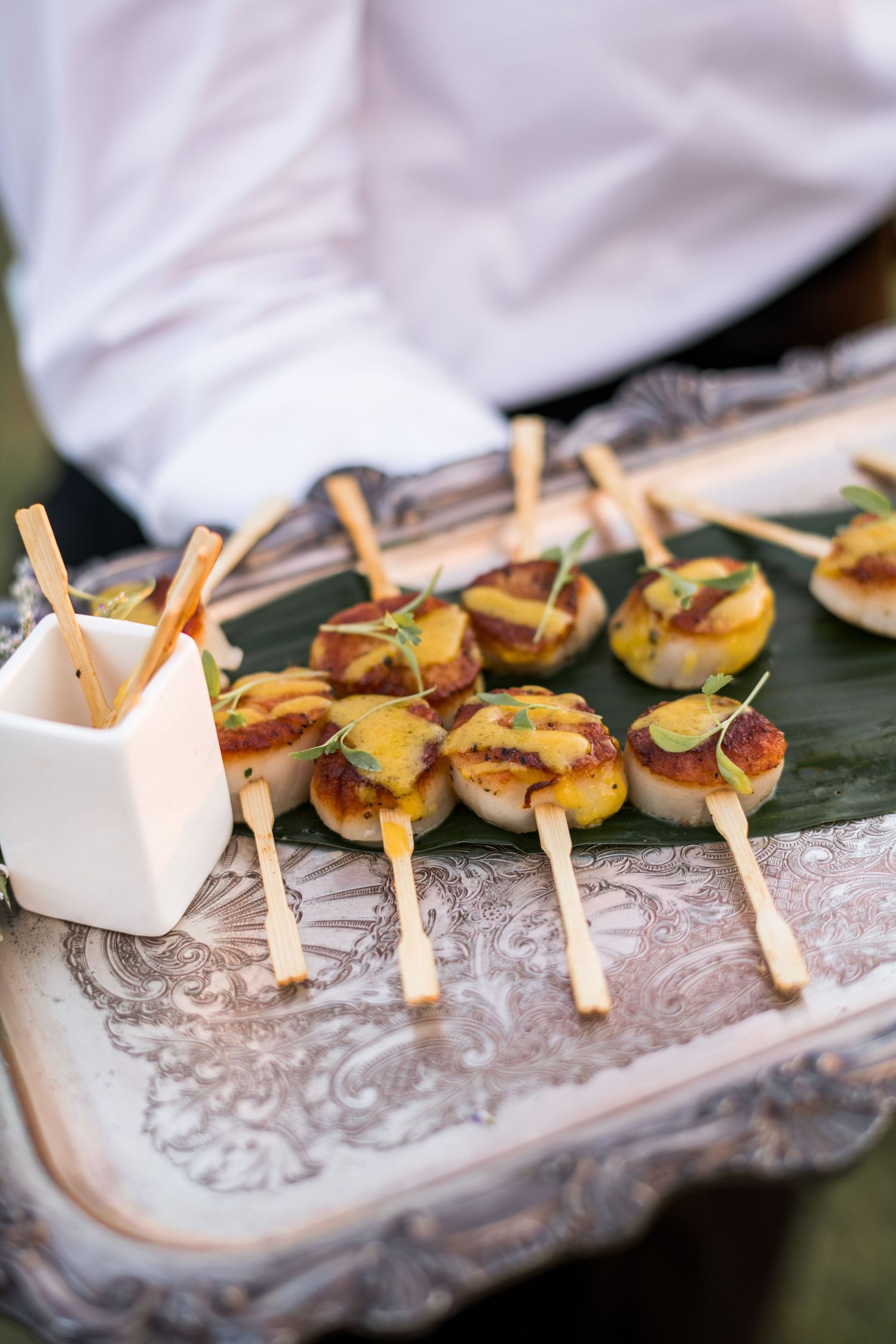 Elegant catering appetizers with grilled scallops on wooden skewers arranged on a decorative silver platter with ice.