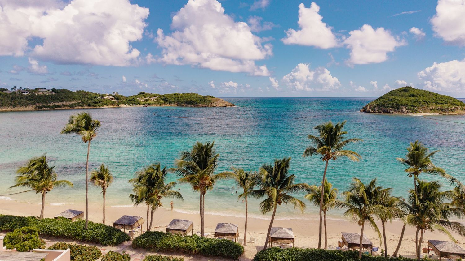 Panoramic view of a tropical beach with palm trees and turquoise waters under blue skies with white clouds.
