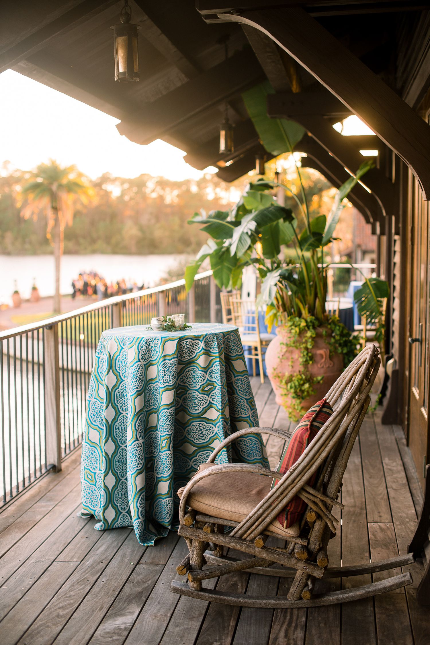 A wooden deck porch with turquoise tablecloths and Adirondack chairs overlooking a waterfront at sunset.